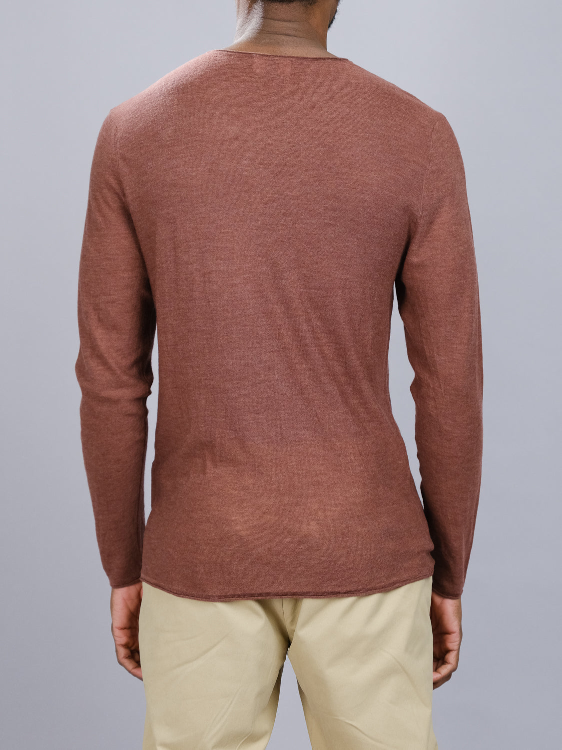 From the back, a person is seen in an exquisitely fitting brown Vogue Lightweight Merino Sweater by ID Menswear and beige pants. The extra fine Merino wools texture contrasts strikingly with the plain gray backdrop, offering a smooth, timeless appeal.