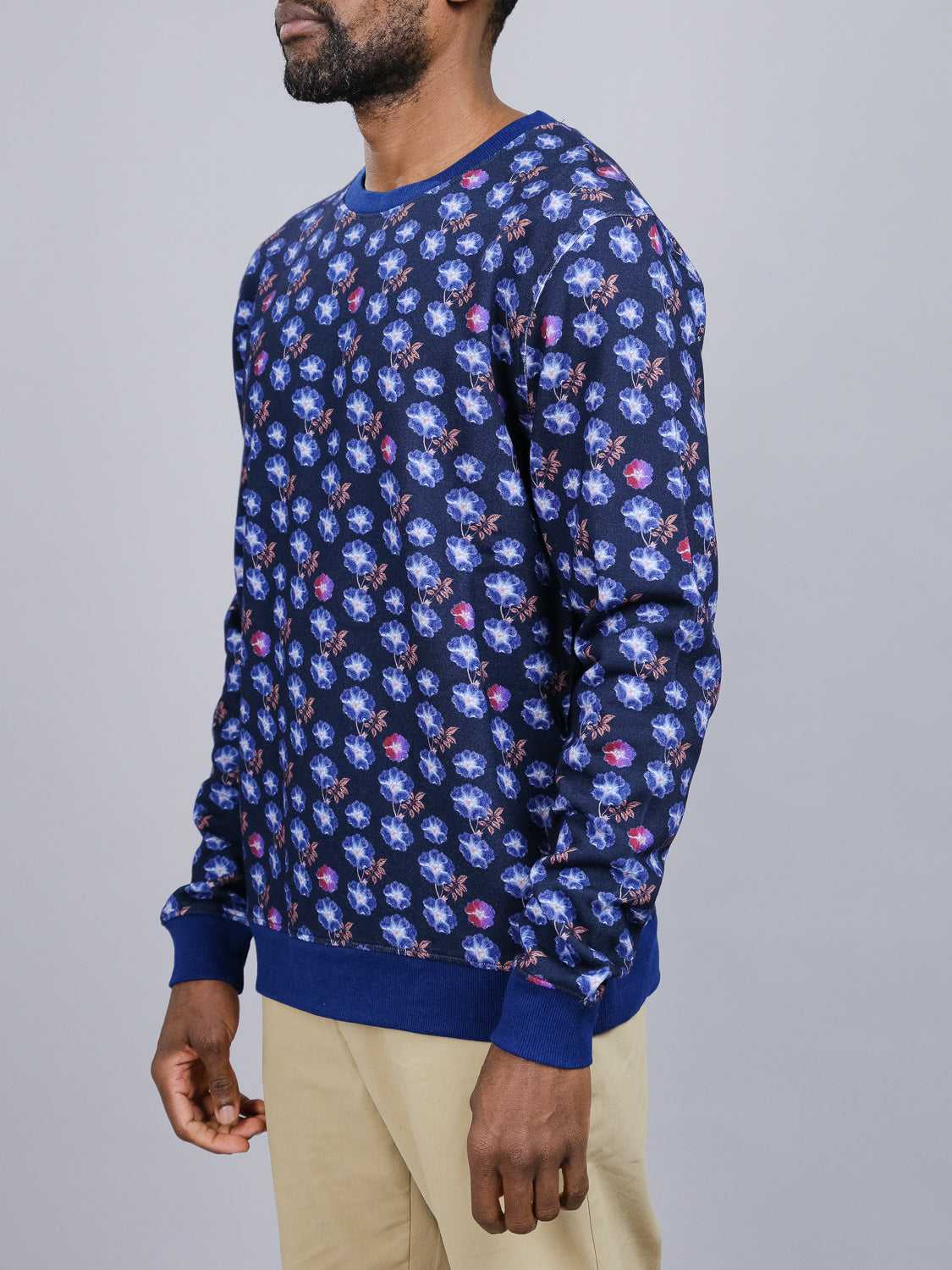 A person in the Flora Printed Sweatshirt by ID Menswear, featuring a vibrant floral digital print made from bamboo and organic cotton fleece, stands against a gray background. Clad in light brown pants with hands relaxed by their sides, the side view highlights the sweatshirts vivid design.