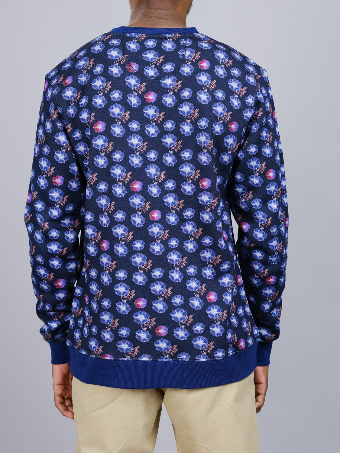 Viewed from the back, a person in khaki pants wears ID Menswears Flora Printed Sweatshirt. It features skull and flower patterns in blue, pink, and purple on bamboo and organic cotton fleece.