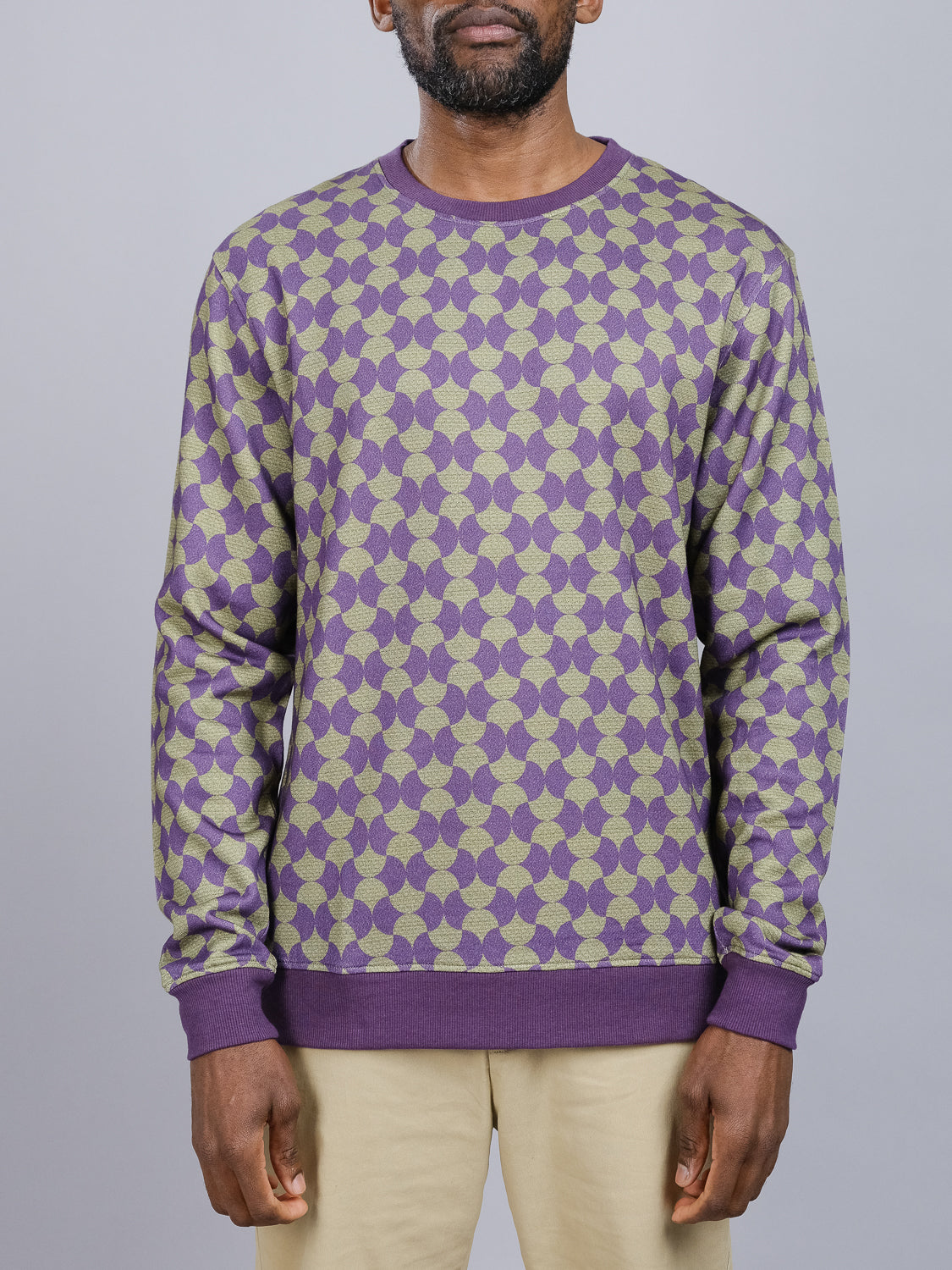 A man poses against a light gray background wearing the Wiggle Geometric Printed Sweatshirt by ID Menswear, showcasing purple and beige patterns with matching cuffs and hem, paired with light pants made from cozy bamboo cotton fleece.
