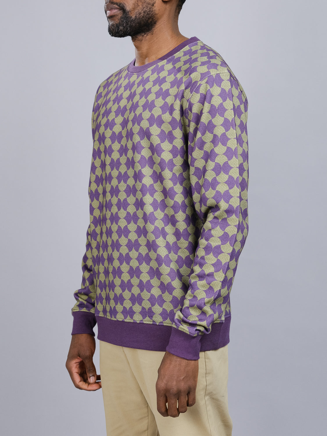 The individual sports an ID Menswear Wiggle Geometric Printed Sweatshirt, showcasing a purple and beige pattern with ribbed cuffs and hem. The look is completed with beige pants against a plain gray backdrop for a casual style.