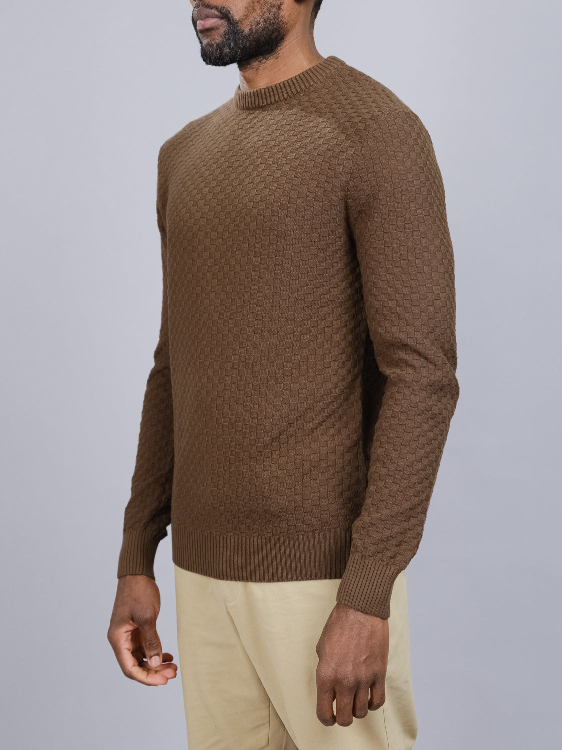 A man in a Turku Cotton Sweater by ID Menswear, featuring a waffle texture and ribbed cuffs, pairs it with beige pants against a gray background for comfort and style.