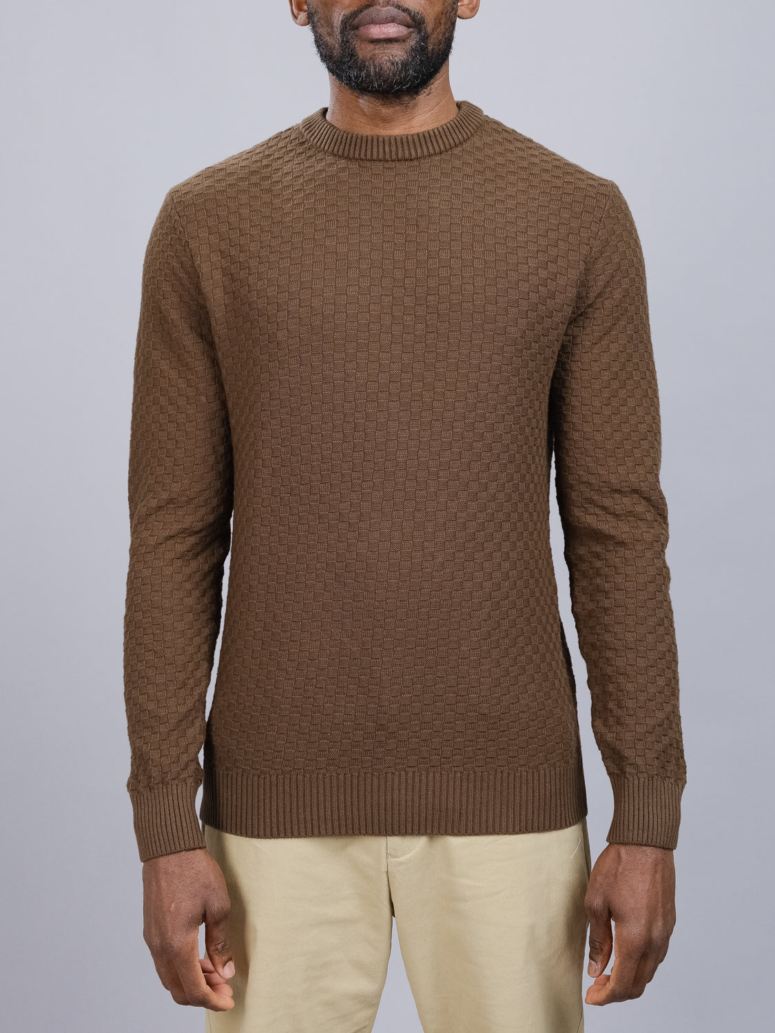 A person wearing the stylish Turku Cotton Sweater by ID Menswear stands against a plain gray background. The focus is on the sophisticated knit design of the sweater, effortlessly paired with beige pants to highlight its elegant texture.