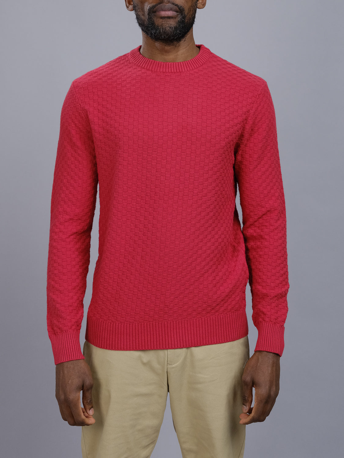A man in an ID Menswear Turku Cotton Sweater and beige pants stands against a gray backdrop. The stylish knit red sweater, woven with a regular fit, is well-lit, showcasing its details.