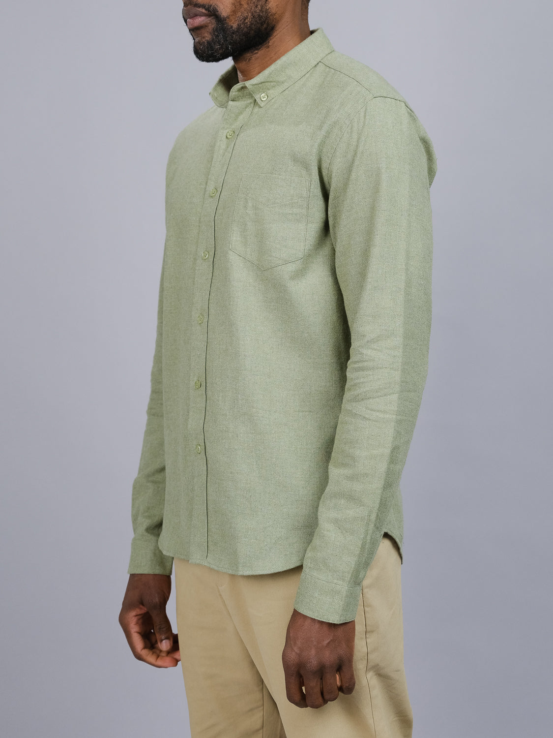 A person in a light green Max Brushed Flannel Shirt by ID Menswear, crafted from 100% cotton with a chest pocket, and beige pants stands against a plain gray background, facing slightly sideways.