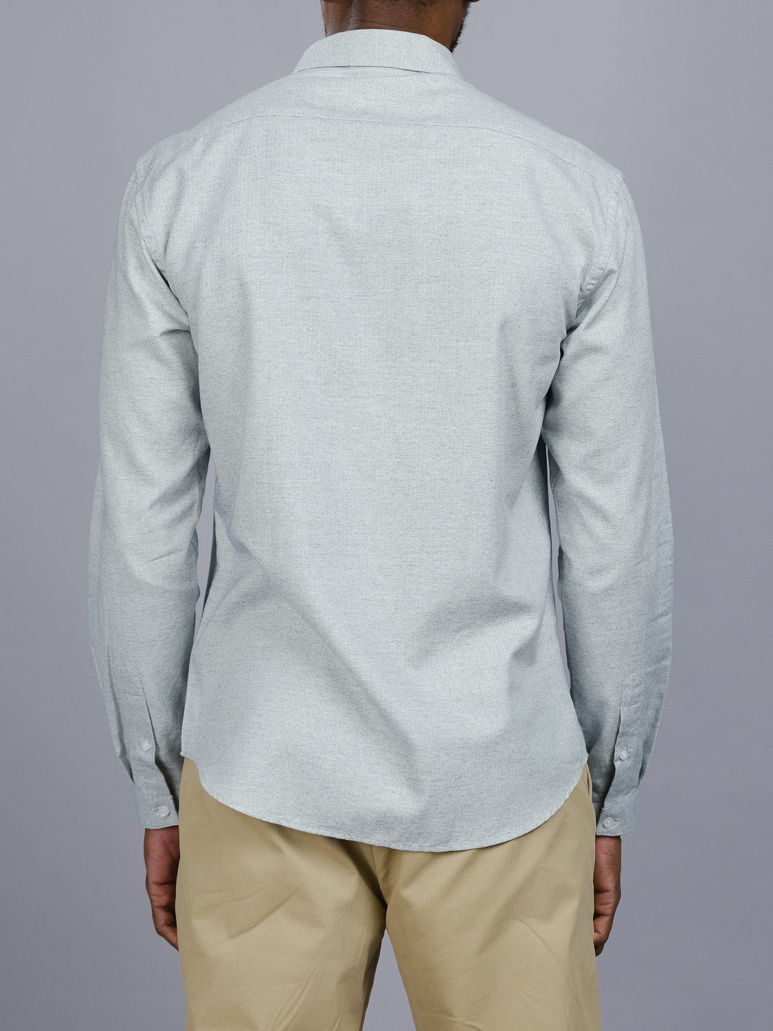 A person is seen from the back wearing a light gray Maya Fine Herringbone Shirt by ID Menswear, paired with beige pants against a plain gray backdrop. This 100% cotton, untucked button-down shirt enhances their relaxed posture with arms at their sides.
