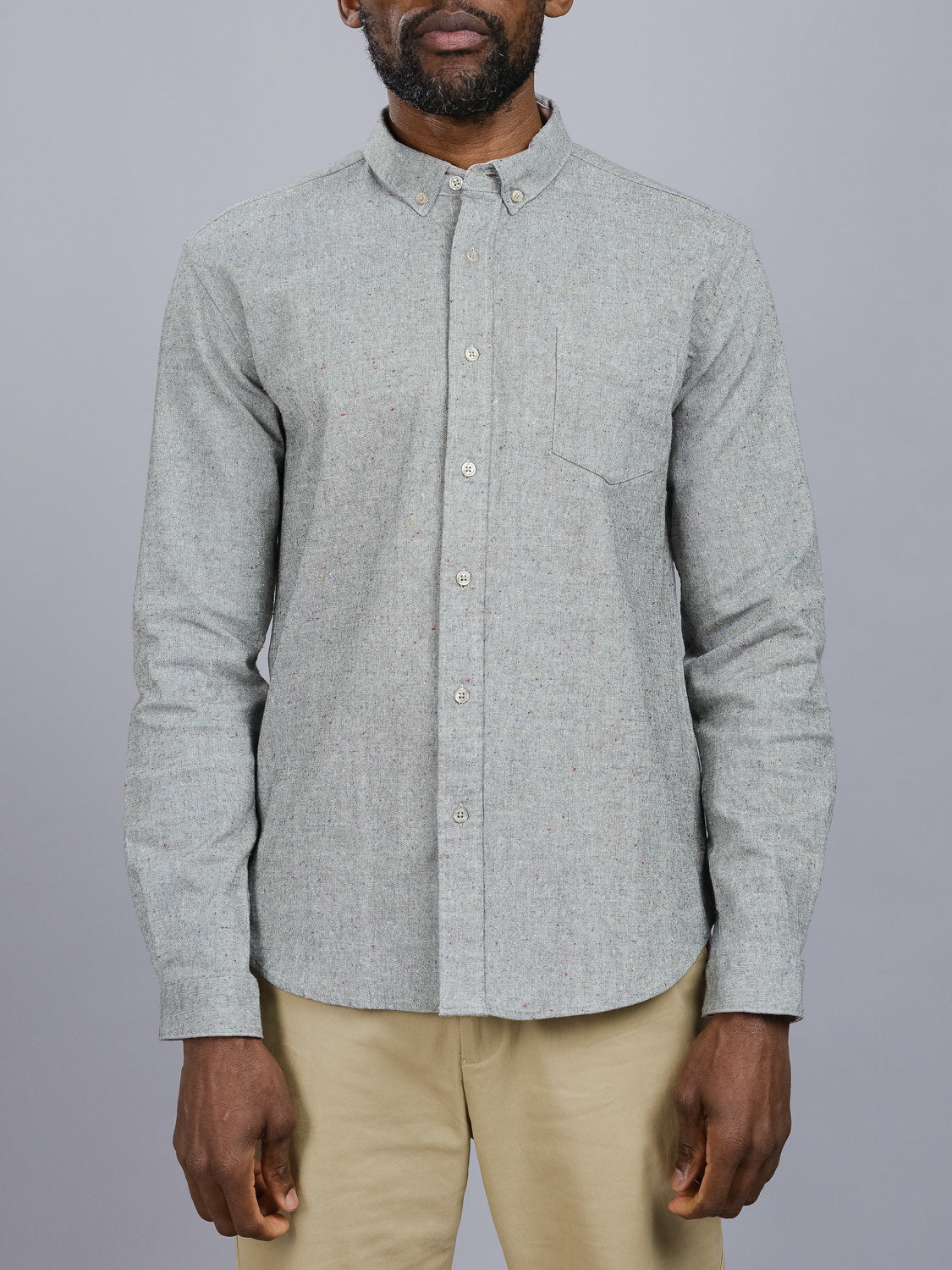 A person wearing an ID Menswear Allswell Speckled Button Down Shirt in premium cotton-linen with a pocket stands against a gray background, paired with beige pants. The shirt’s subtle texture and sleeves are rolled down, highlighting its versatile design.