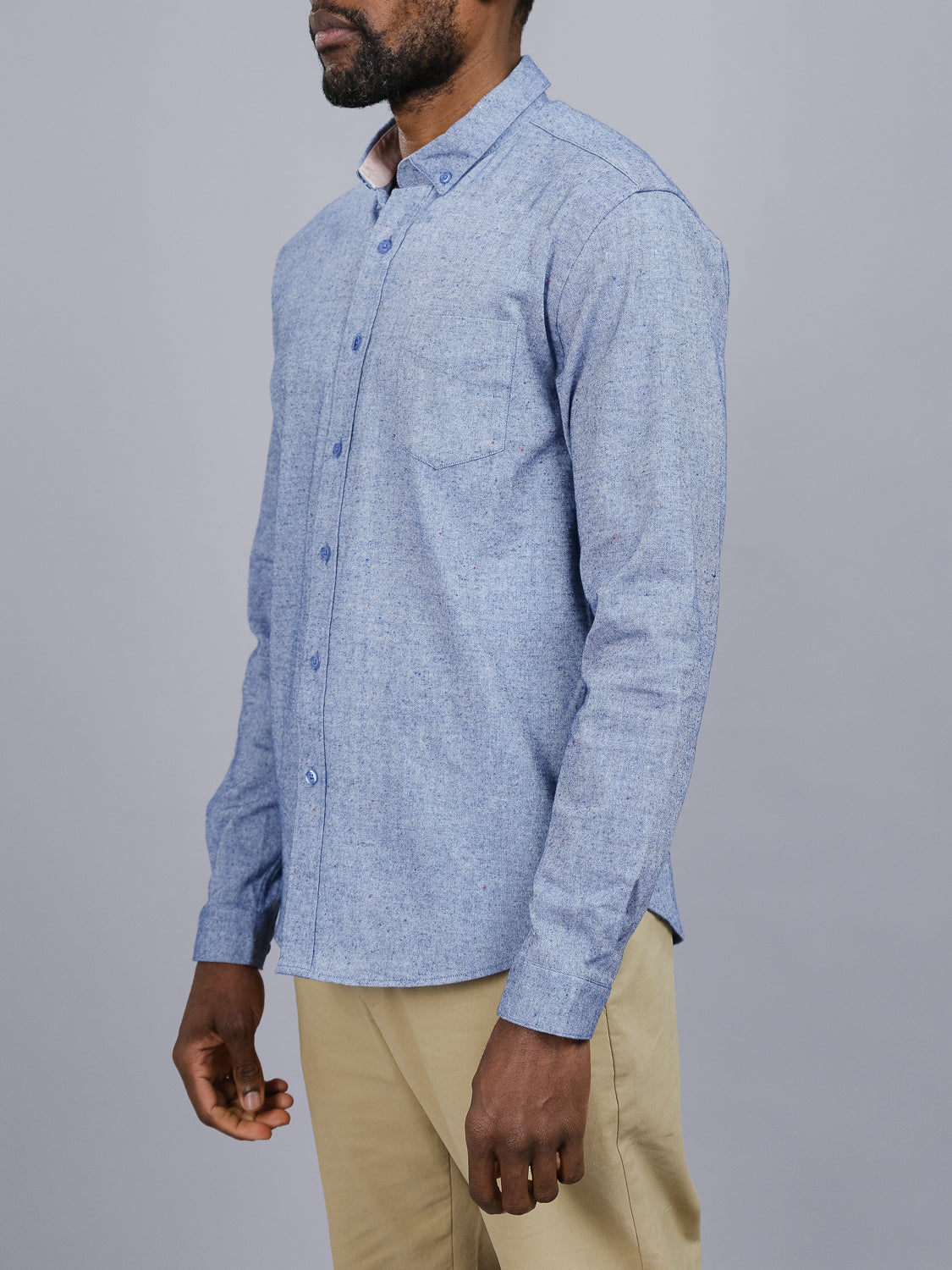 Clad in ID Menswears Allswell Speckled Button Down Shirt, a versatile light blue shirt made from premium cotton-linen with a chest pocket, and beige pants, the person is photographed side profile against a plain gray backdrop.