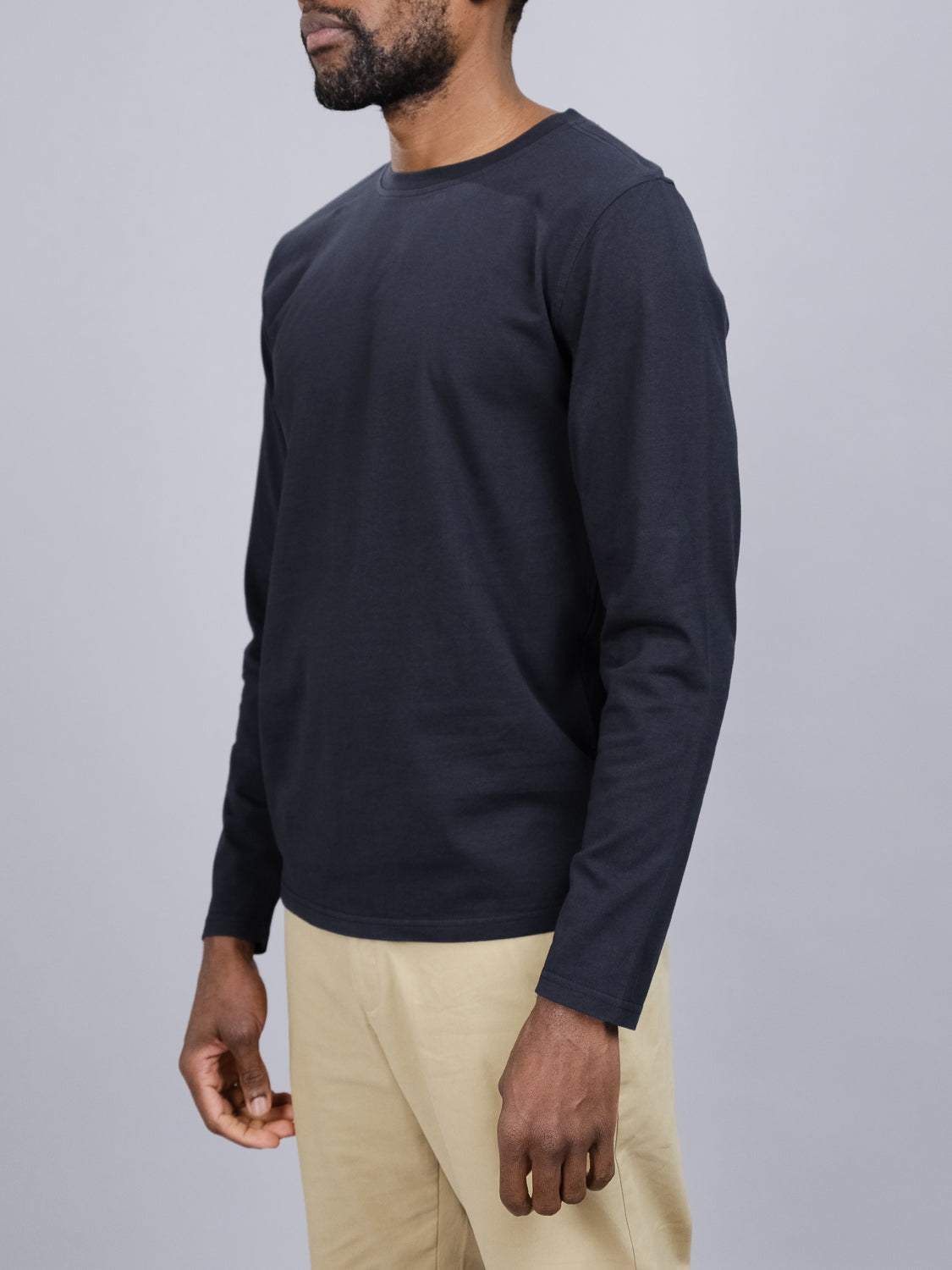 A person models the Sublime Long Sleeve Tee by ID Menswear, crafted from organic cotton in black, paired with beige pants. They stand against a gray background, shown from the shoulders down, highlighting the casual style.