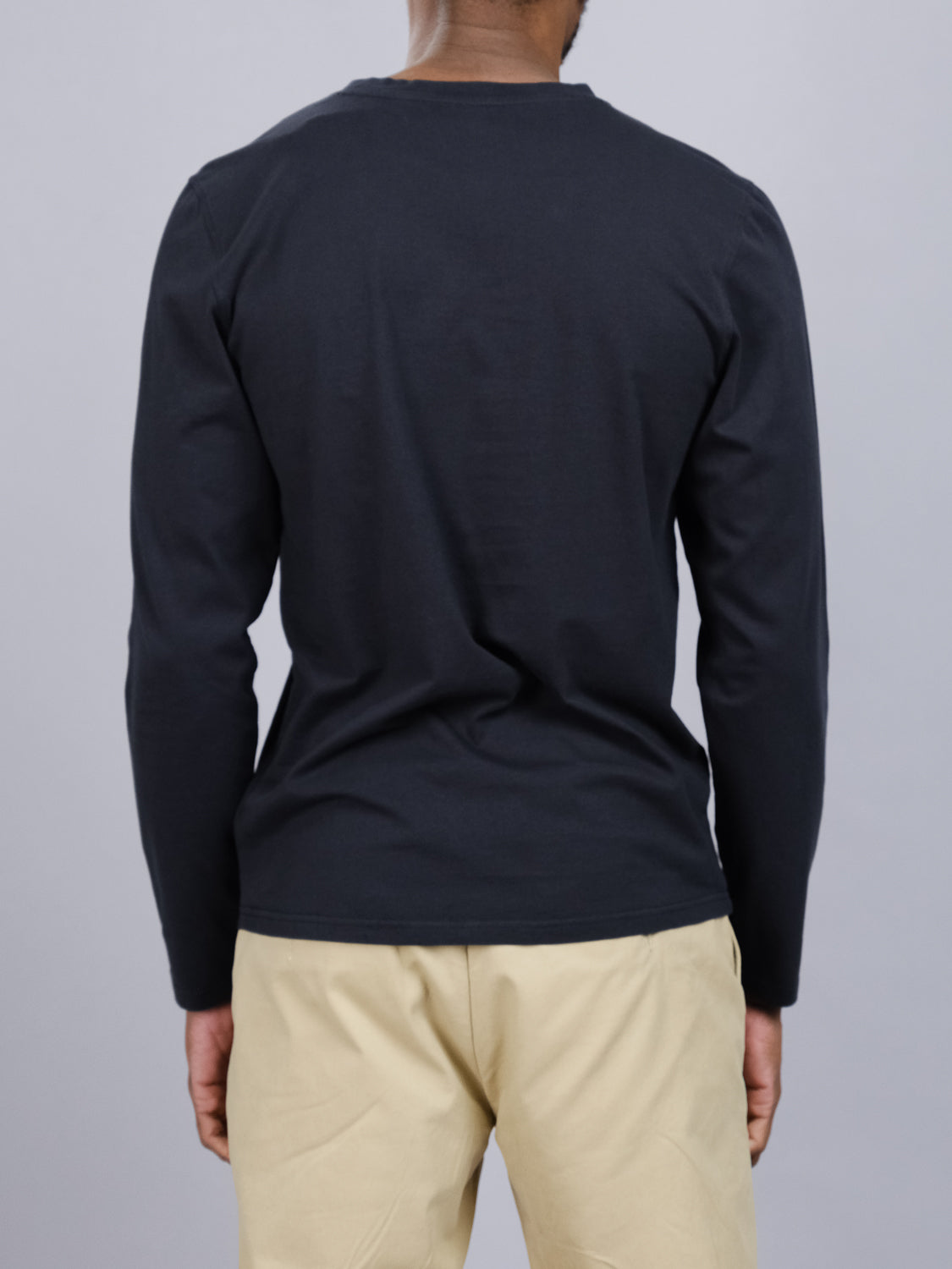 Someone is seen from behind wearing the ID Menswear Sublime Long Sleeve Tee in organic cotton, black, paired with beige pants, set against a gray backdrop.