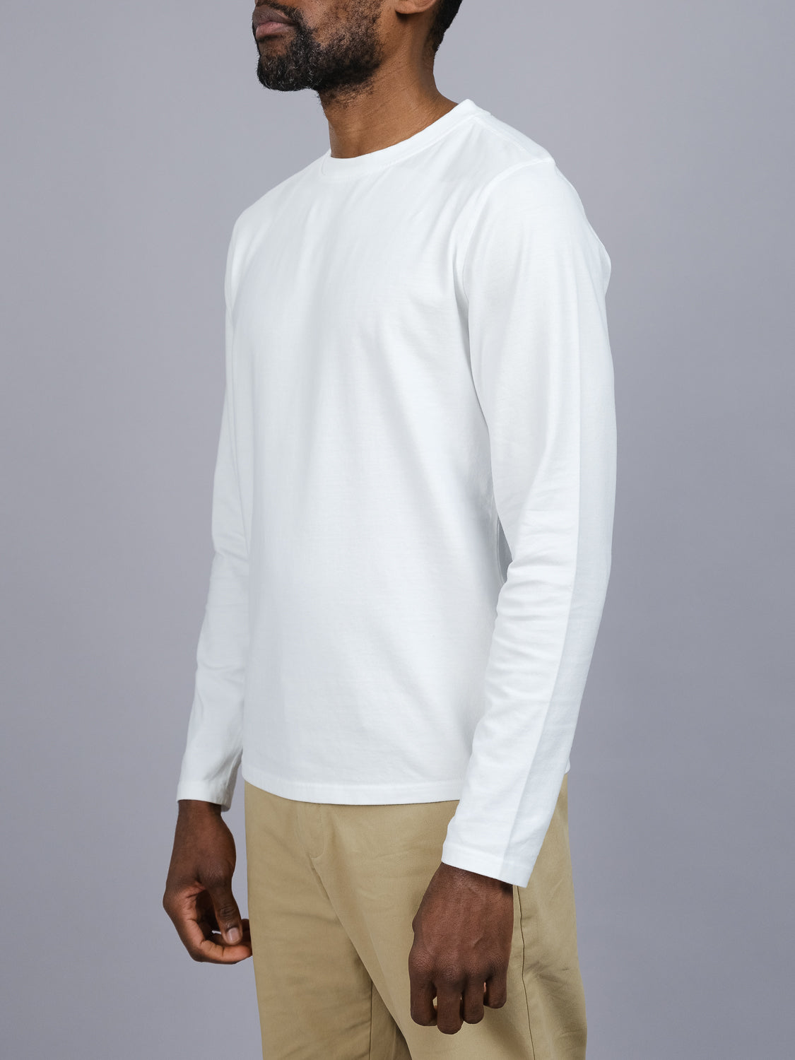 A man models the ID Menswear Sublime Long Sleeve Tee, crafted from organic cotton in plain white, paired with beige pants. He faces slightly right against a gray backdrop, showcasing his left arm and resting his right hand casually at his side for an effortless look.