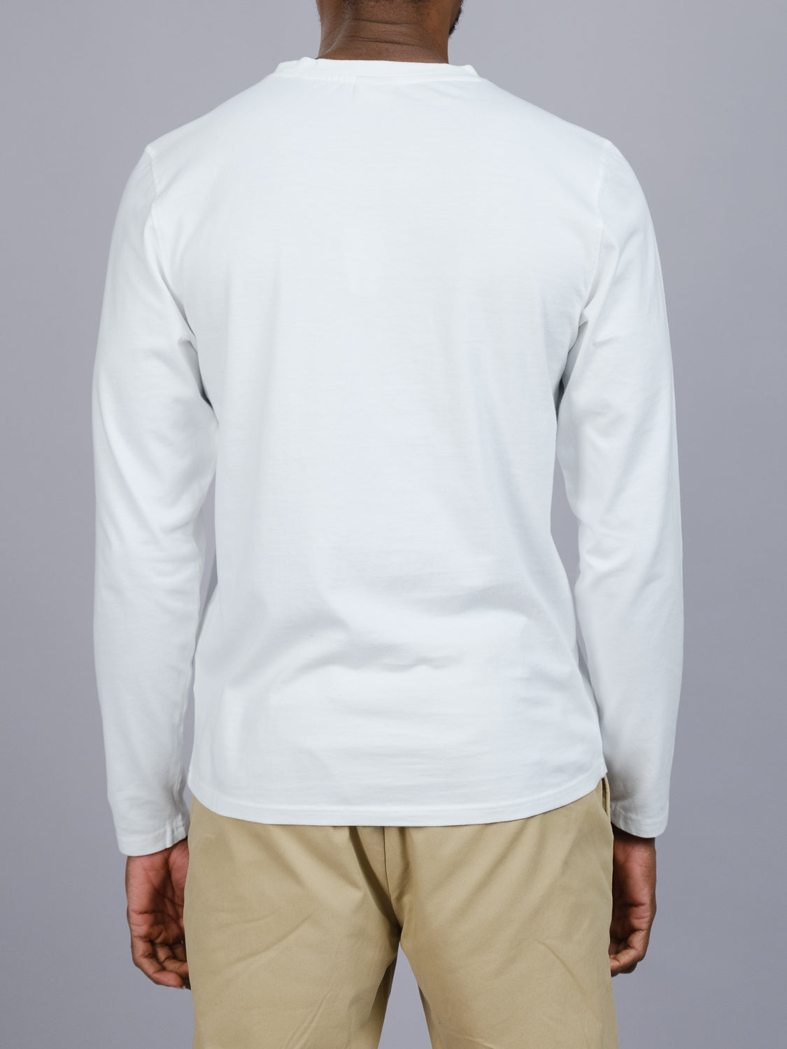 A person is standing with their back to the camera against a plain gray background, wearing ID Menswears light gray Sublime Long Sleeve Tee and beige pants made from organic cotton.