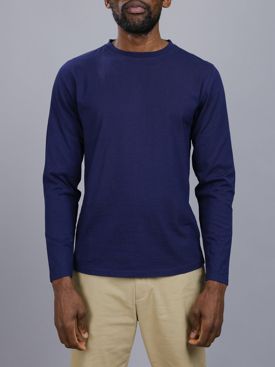 Amidst a gray backdrop, an individual models ID Menswears Sublime Long Sleeve Tee in navy blue organic cotton and beige pants. They maintain a neutral expression with hands casually at their sides.