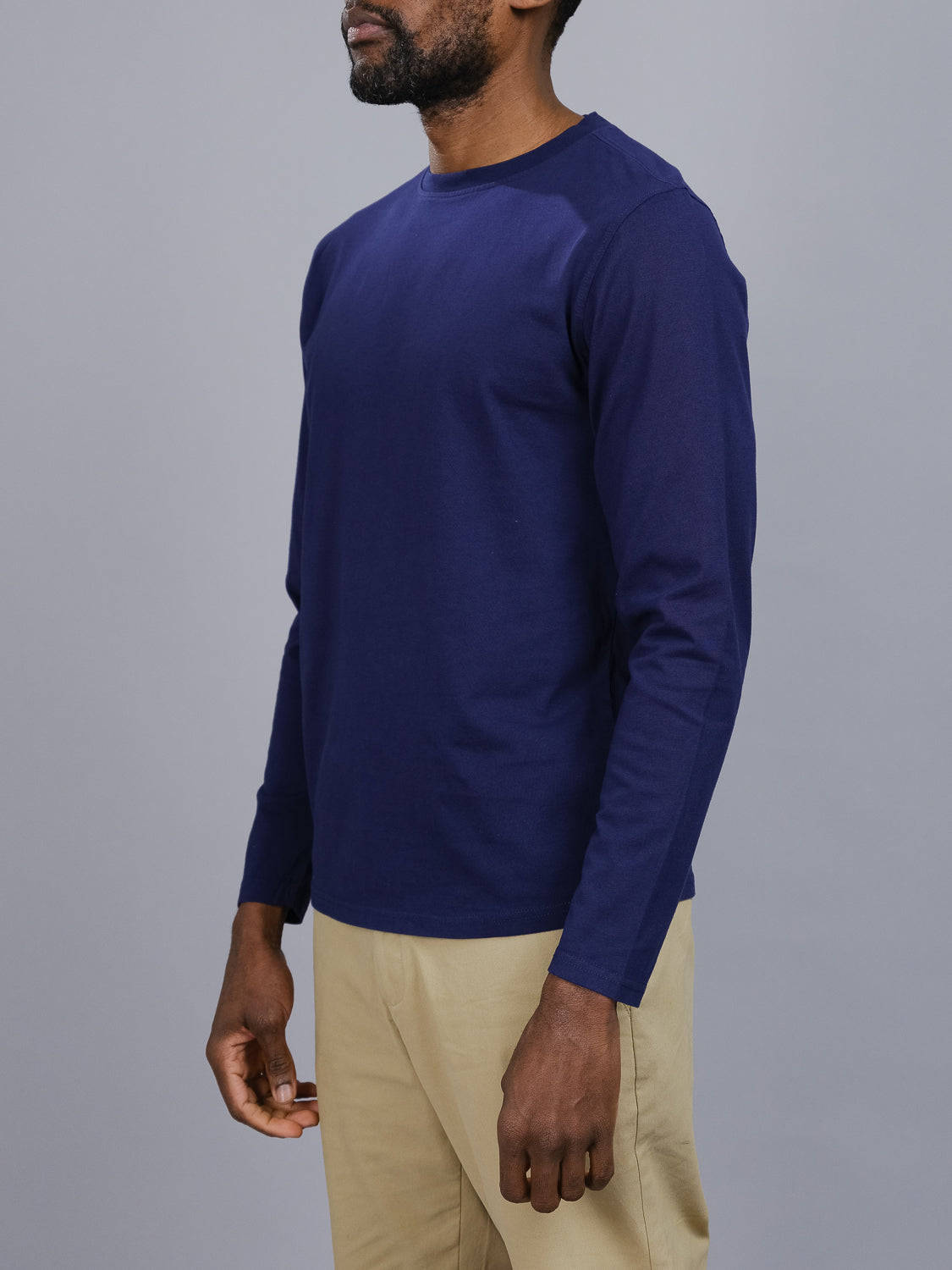 A person models the Sublime Long Sleeve Tee from ID Menswear, featuring a navy blue hue and crafted from organic cotton. They pair it with beige pants against a plain gray background, not showing their face, encapsulating casual style.