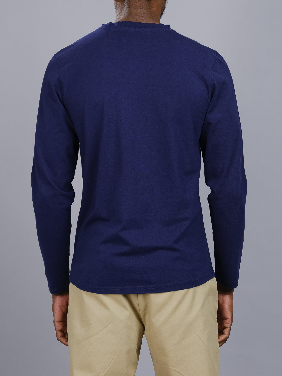An individual in a Sublime Long Sleeve Tee from ID Menswear, featuring navy blue organic cotton, and beige pants stands casually with their back to the camera against a gray background.