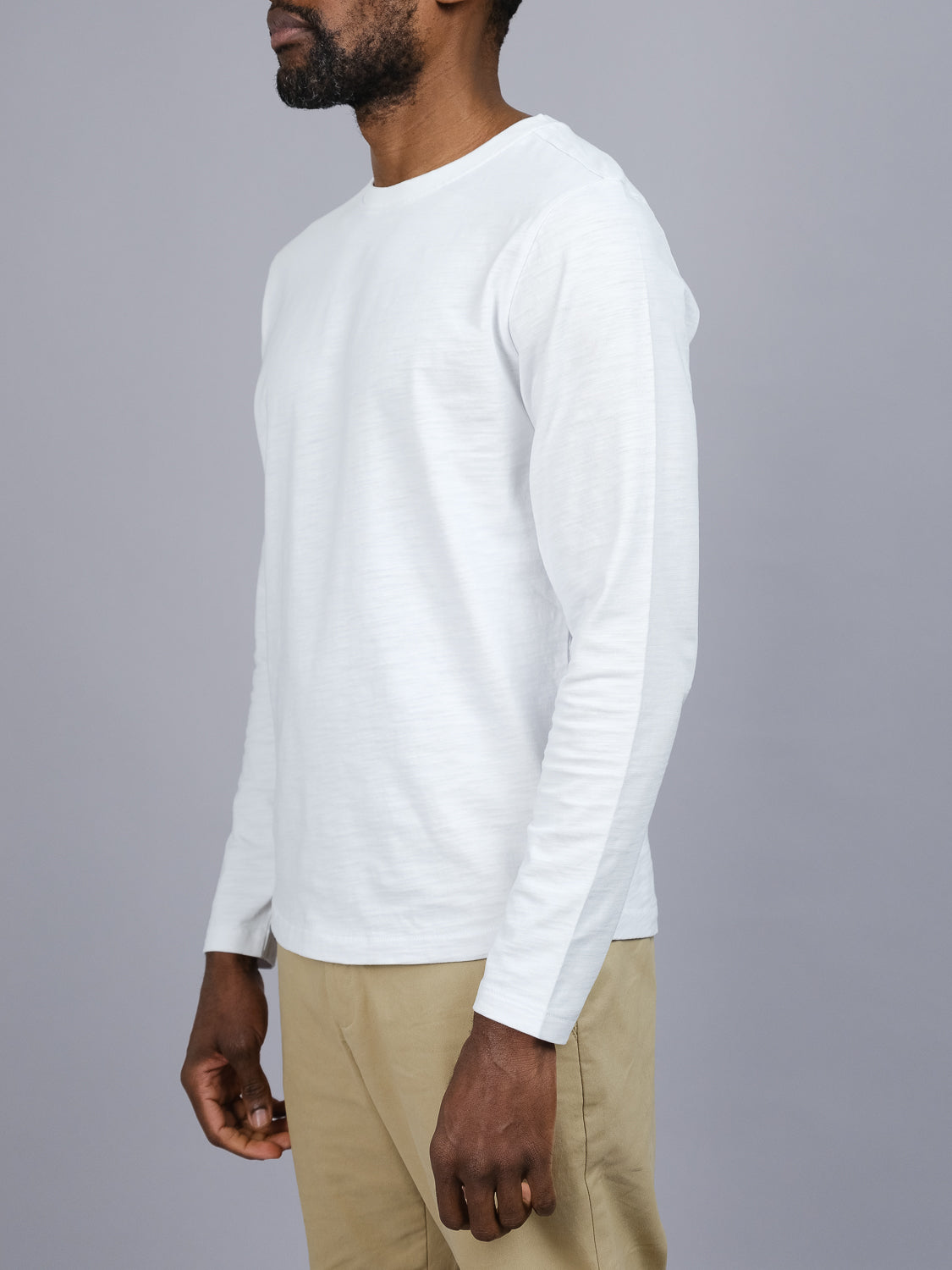 A man in the ID Menswear Paradise Long Sleeve Tee in plain white cotton and khaki pants stands against a gray background, his head slightly turned to the side, epitomizing effortless casual wear.