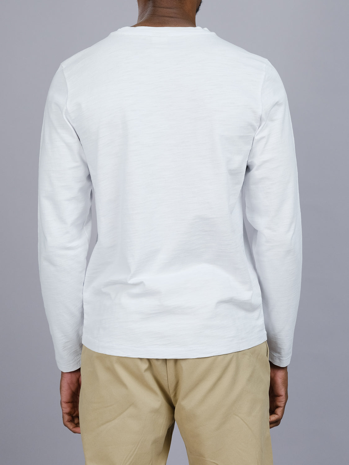 A person in a white Paradise Long Sleeve Tee by ID Menswear and beige pants stands with their back to the camera against a gray backdrop, showcasing effortless casual wear.