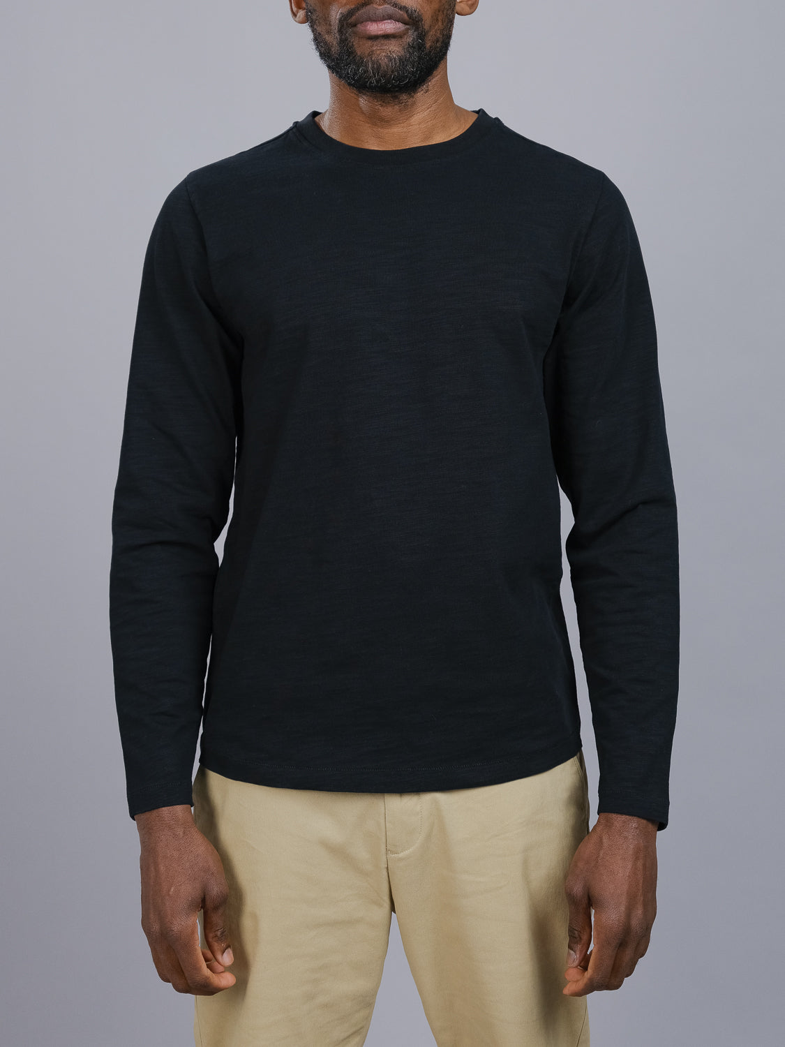 A person in a Paradise Long Sleeve Tee by ID Menswear and beige pants stands against a gray background. Their relaxed arms highlight the casual ensemble, with the slub cotton fabric adding effortless texture.