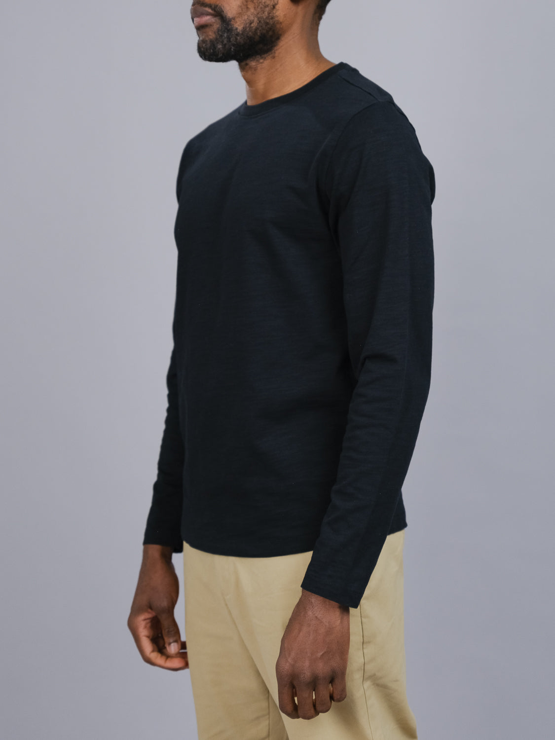 Sporting ID Menswears Paradise Long Sleeve Tee in black and beige pants, this individual stands against a gray backdrop. They strike a casual pose with relaxed arms, slightly turned to the side, effortlessly showcasing their style.