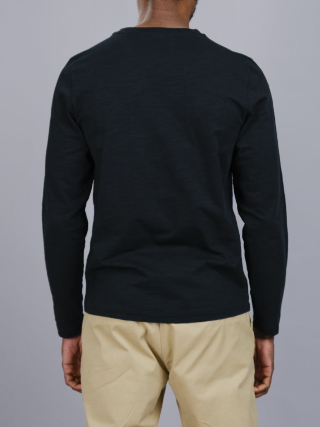 Wearing ID Menswears black Paradise Long Sleeve Tee and beige pants, a person stands with their back to the camera against a plain gray background, epitomizing effortless casual style.