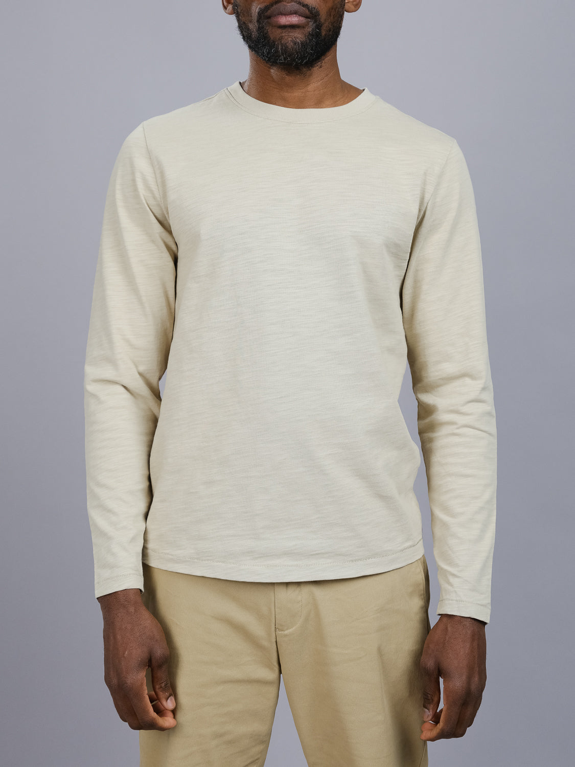 A person models the Paradise Long Sleeve Tee by ID Menswear, crafted in light beige slub cotton, paired with tan pants against a neutral gray backdrop. The emphasis is on the casual attire, leaving the models face out of view.
