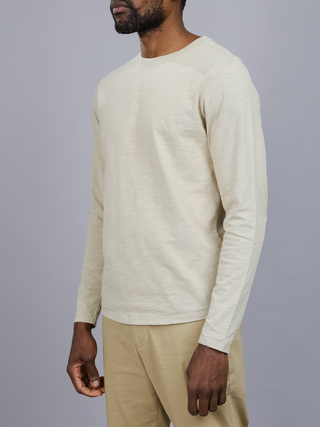 A person in a light beige Paradise Long Sleeve Tee by ID Menswear and khaki pants stands against a gray background, with the shirts slub cotton texture adding sophistication to their casual look as they pose with one arm relaxed by their side.