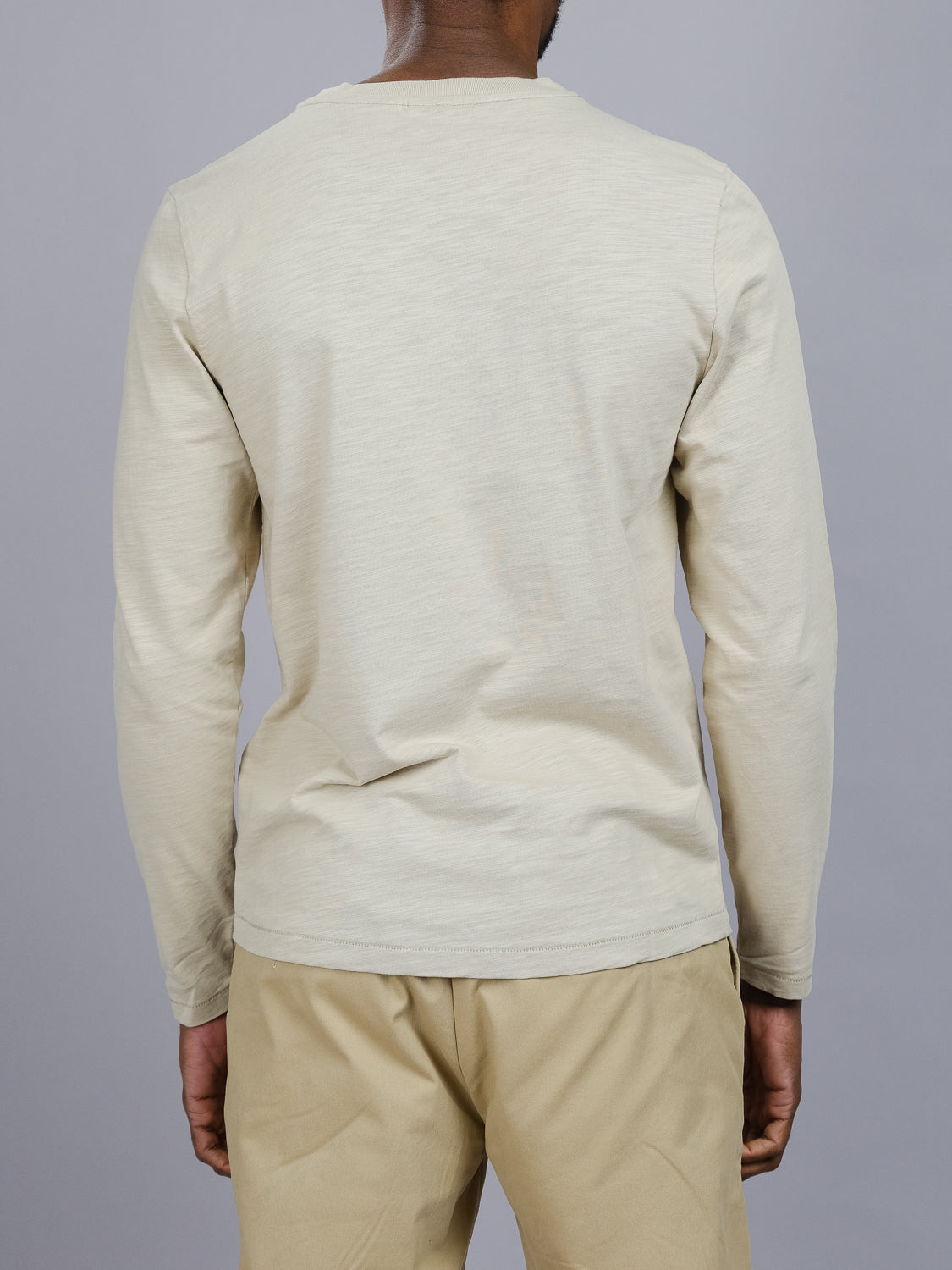 Rear view of a person dressed in ID Menswears Paradise Long Sleeve Tee, light beige, standing against a plain gray background. The focus is on the slub cotton attire from the back.