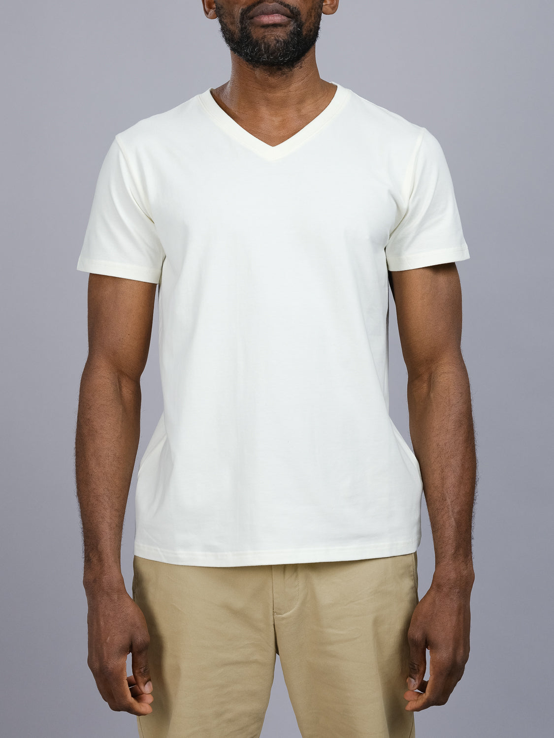 Against a gray background, a person wears ID Menswears Classic V Neck Tee in white, made from a comfy cotton-spandex blend, paired with beige pants. The focus is on the upper body and clothing essentials, with their arms relaxed at their sides.