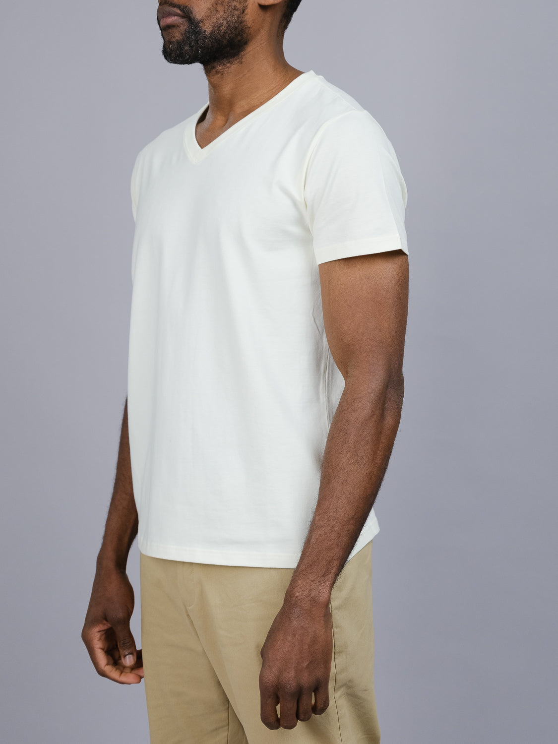 A man wears an ID Menswear Classic V Neck Tee, a wardrobe essential in a comfortable cotton-spandex blend, paired with beige pants. He stands against a plain gray background with his arms by his sides.
