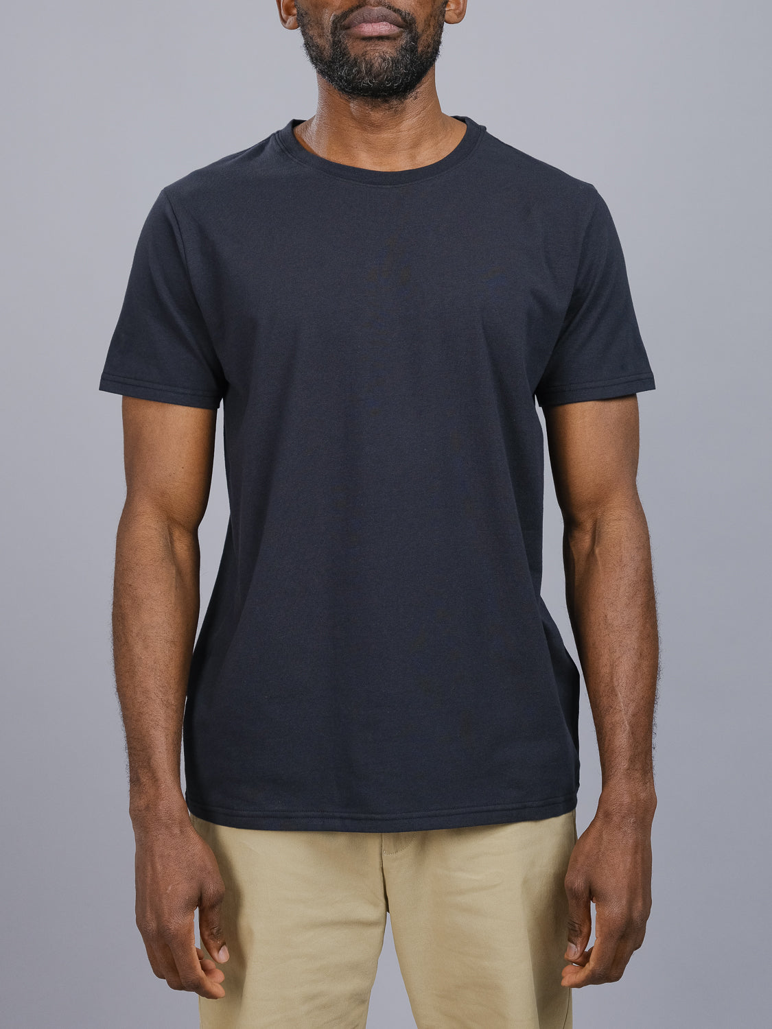 A person stands against a gray background wearing an ID Menswear Classic Crew Neck Tee in plain black, paired with beige pants. The cotton-spandex shirt offers a regular fit, and their arms are relaxed by their sides.