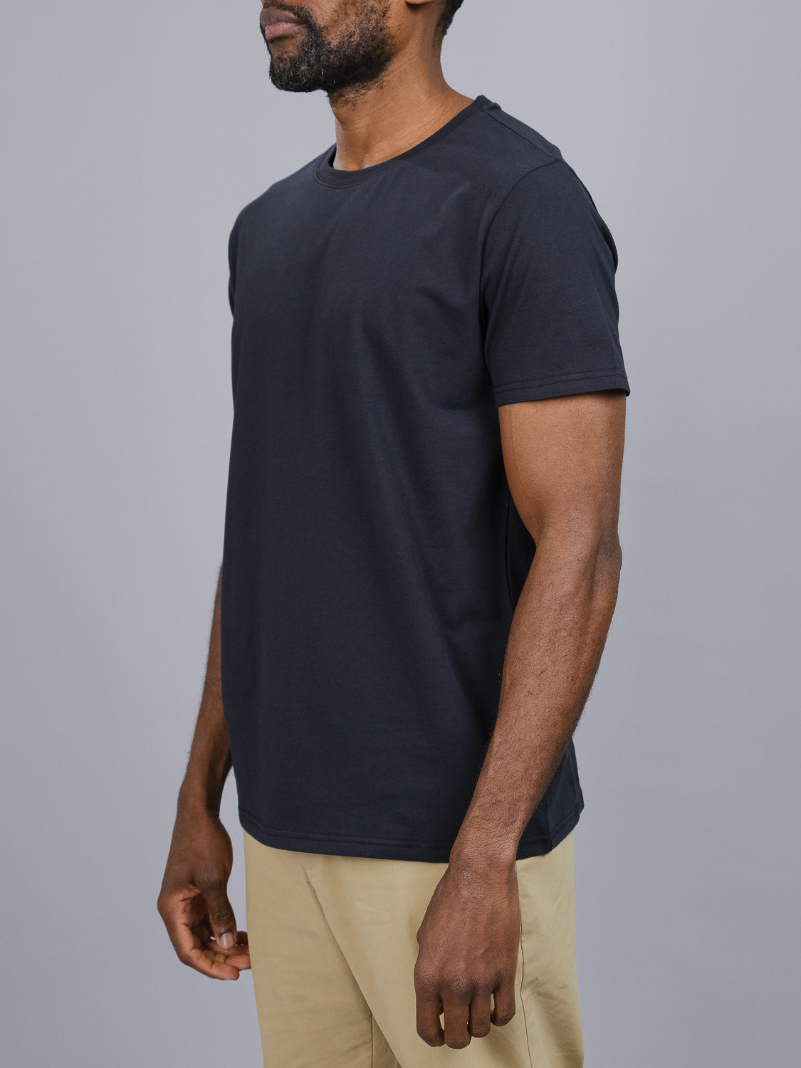 A person stands against a gray background wearing an ID Menswear Classic Crew Neck Tee in black, paired with beige pants. The cotton-spandex blend ensures a comfortable regular fit, with their arms resting at their sides. The focus is on the outfit, as the photo crops out the face above the nose.