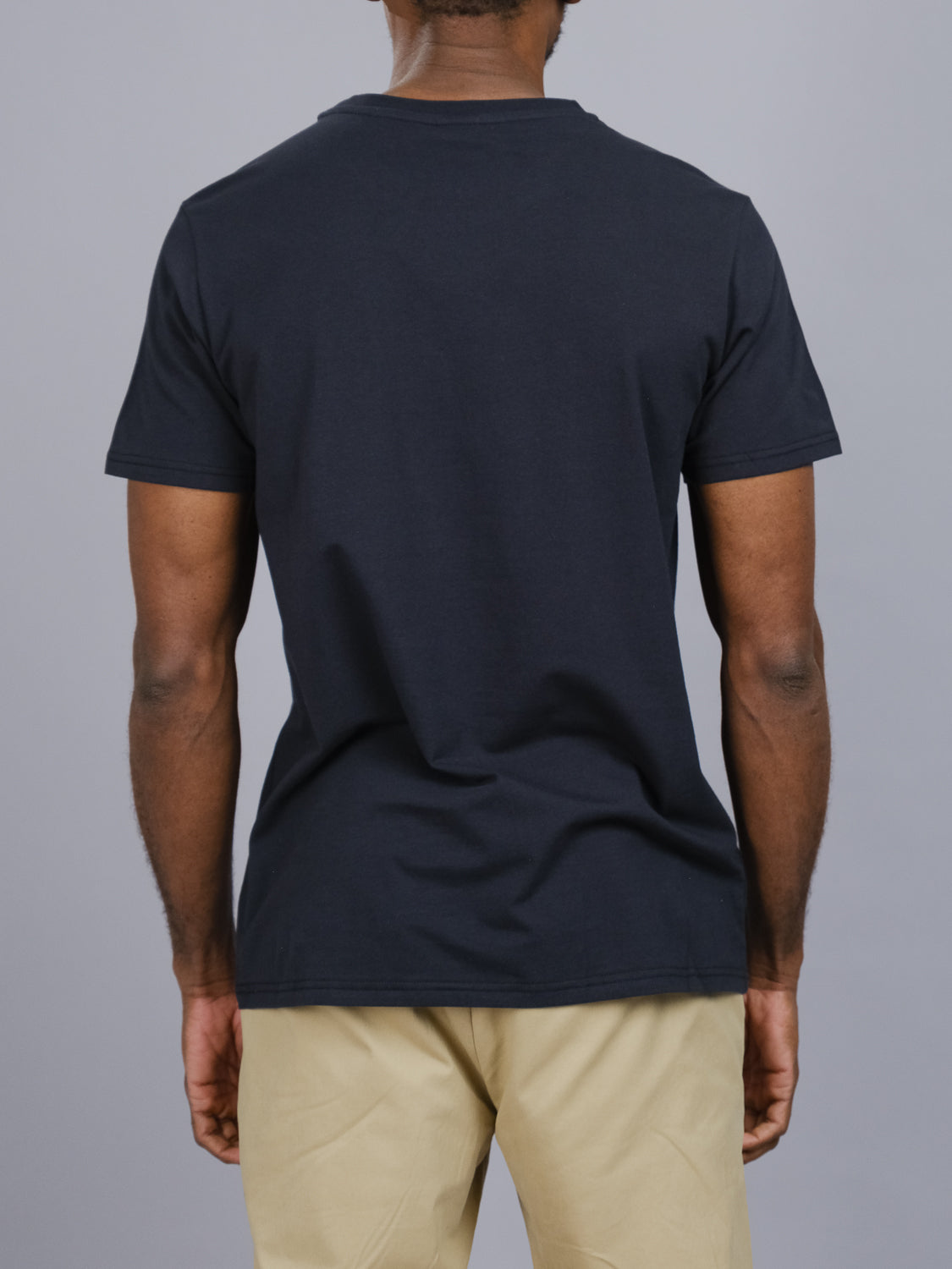 An individual in an ID Menswear Classic Crew Neck Tee, made of cotton-spandex, and beige pants is facing away against a gray backdrop.