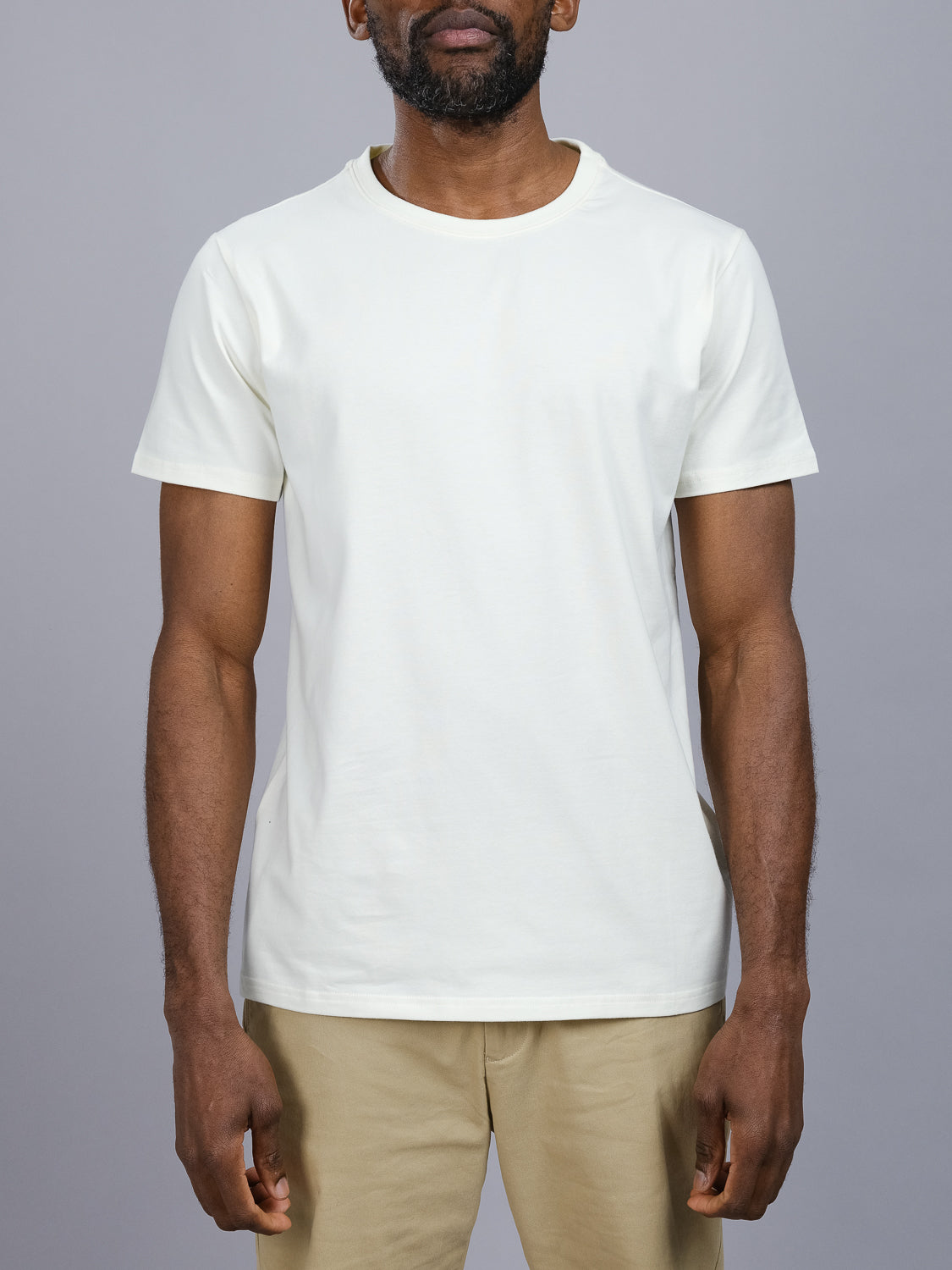 A person wearing an ID Menswear Classic Crew Neck Tee, made of soft cotton-spandex fabric, and beige pants stands against a gray background. The image captures them from the shoulders down in a flattering regular fit with their arms relaxed at their sides.