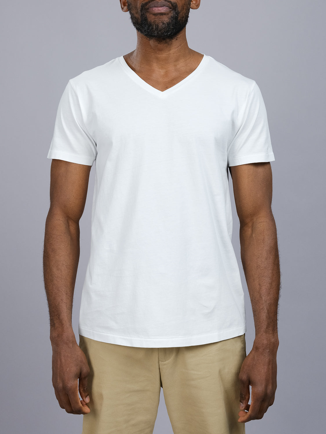 A person in ID Menswears Legacy Organic Cotton V Neck Tee and beige pants stands against a gray background. With short dark hair and a beard, they present a timeless, vintage look.