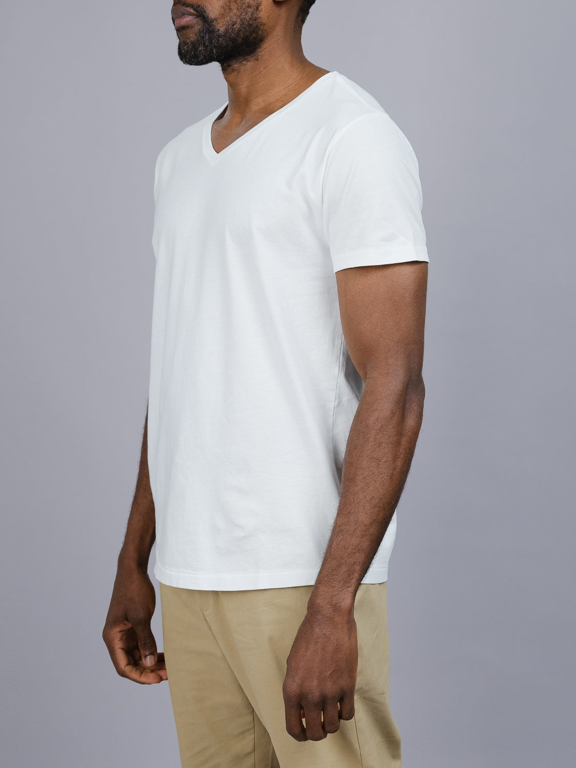 Someone in an ID Menswear Legacy Organic Cotton V Neck Tee and beige pants stands against a plain gray background, upper body partially turned to the side.
