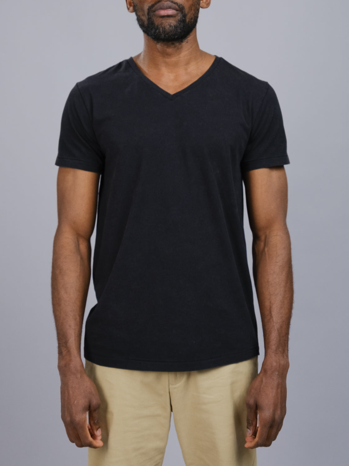 A man models the ID Menswear Legacy Organic Cotton V-Neck Tee in classic fit, plain black, paired with beige organic cotton pants. He stands against a gray background, arms by his sides, with the image cropped from shoulders to mid-thigh.
