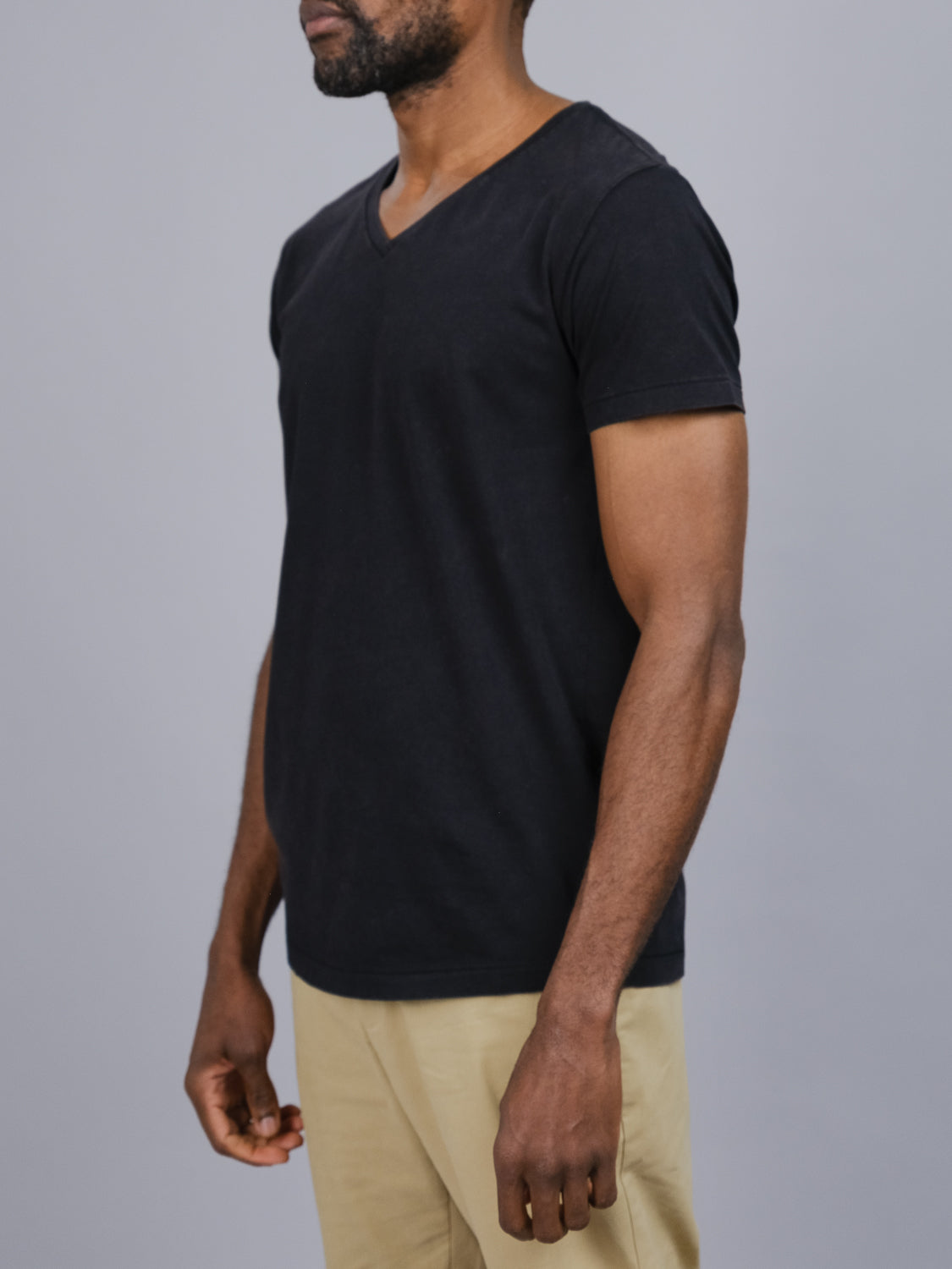 A person in an ID Menswear Legacy Organic Cotton V Neck Tee in black and beige pants stands against a gray background. Captured from the side, the image showcases their upper half with relaxed posture, embodying a classic fit style.