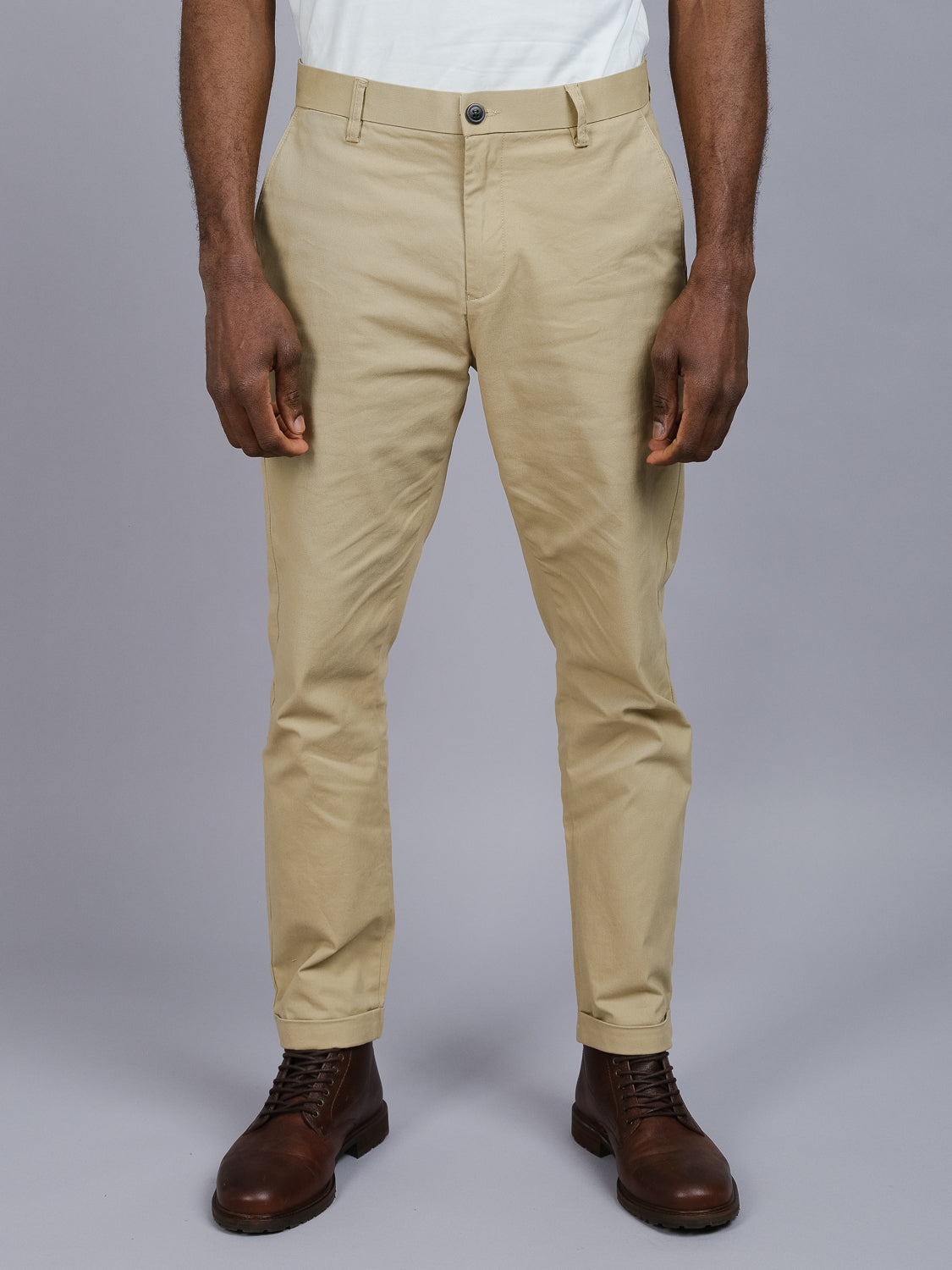 Wearing ID Menswears Java Chino Pant in beige cotton twill and a white shirt, paired with brown leather boots, a person stands against a gray background. The image emphasizes the clothing details from shoulders to feet.