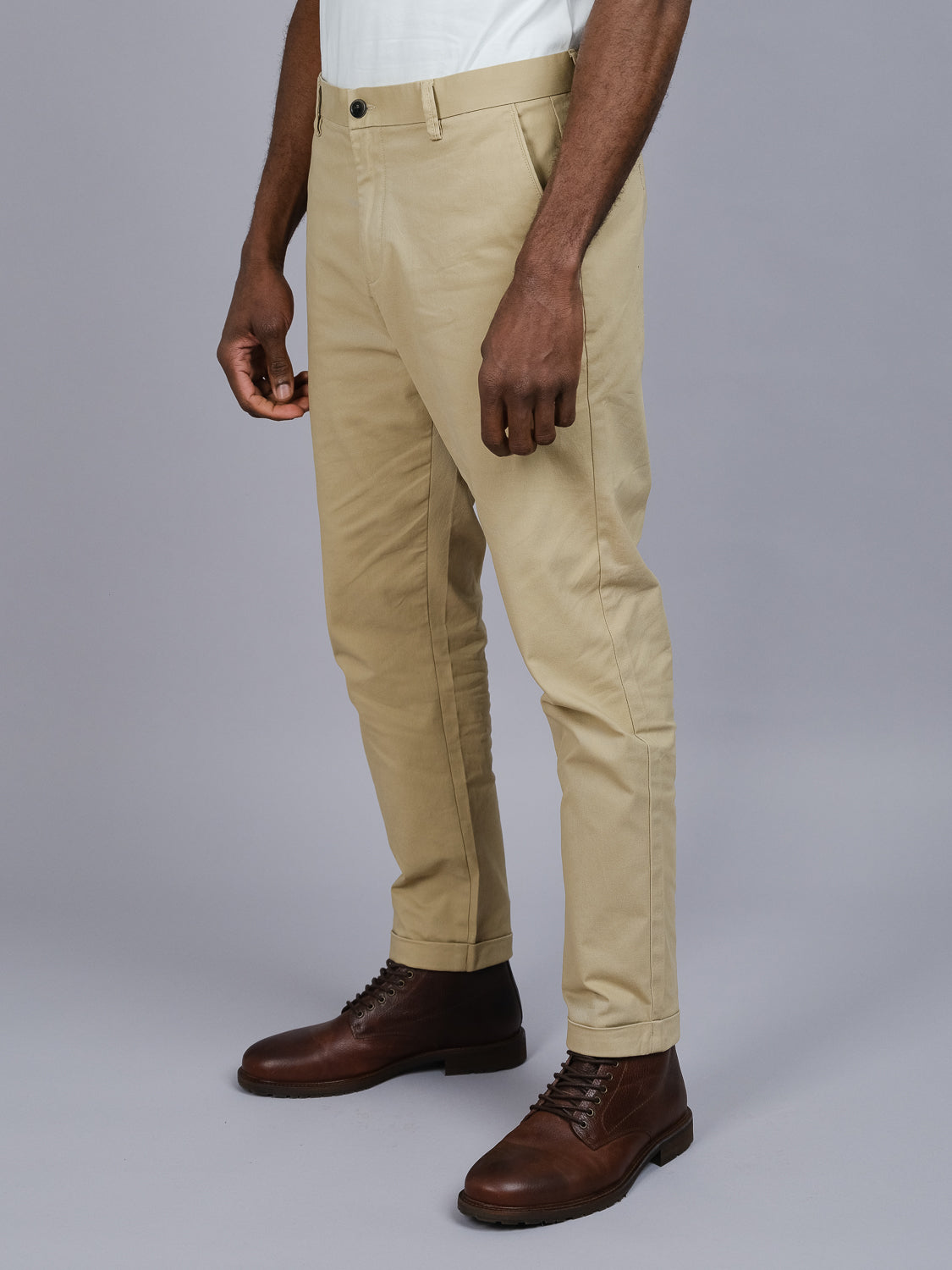 An individual in ID Menswears Java Chino Pant, paired with a white shirt and brown leather boots, stands against a gray background. The focus is on their lower body with arms relaxed at their sides. Their face remains out of the photo.