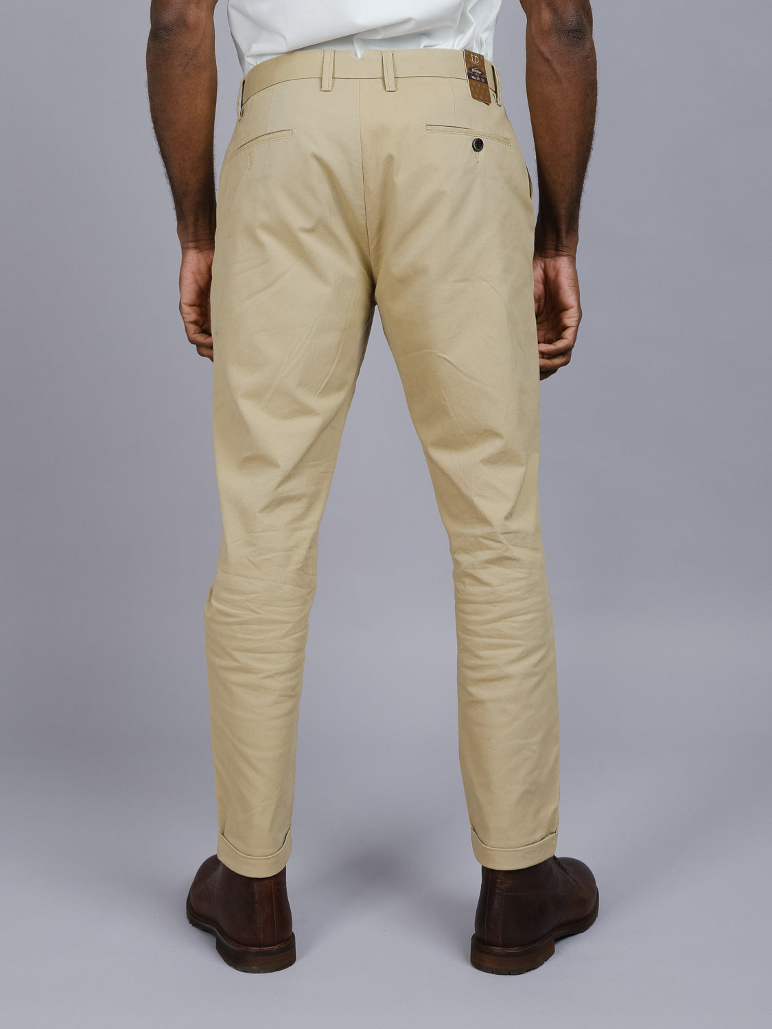 Rear view of a person wearing relaxed fit, beige Java Chino Pant by ID Menswear and brown boots, facing away from the camera. The cotton twill trousers feature pockets and belt loops against a plain grey background.