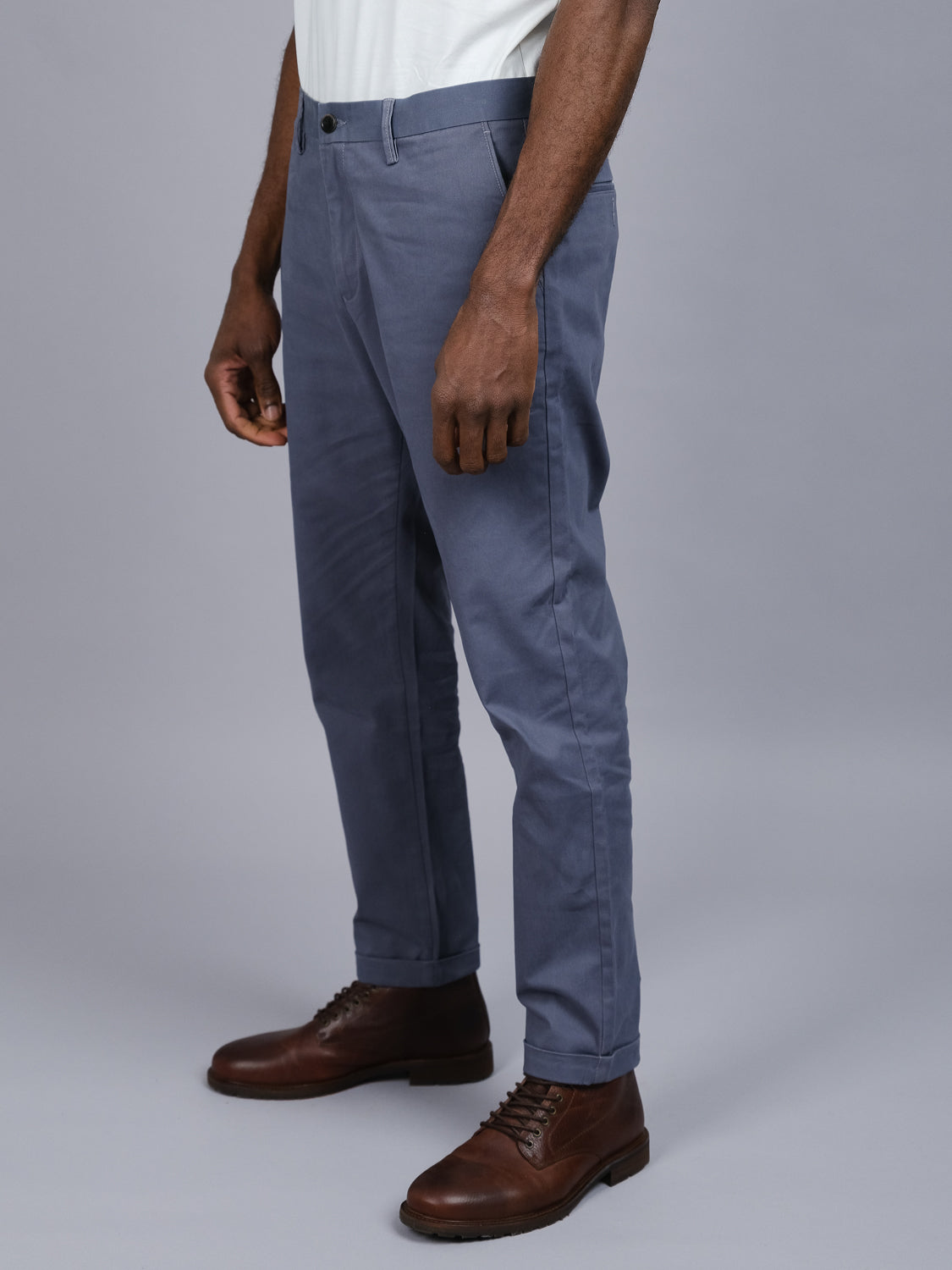 Someone wearing ID Menswears Java Chino Pant in relaxed-fit gray cotton twill stands with one hand along their leg, paired with brown leather shoes against a neutral gray background.