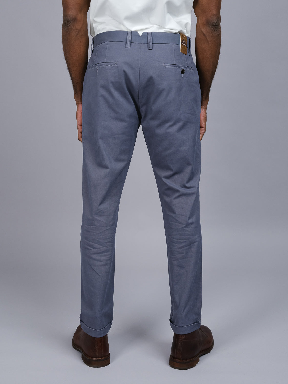A person stands in ID Menswears Java Chino Pant—relaxed fit, blue cotton twill trousers with a waistband brown patch—and a white shirt. Brown shoes complete the look against a plain gray backdrop.