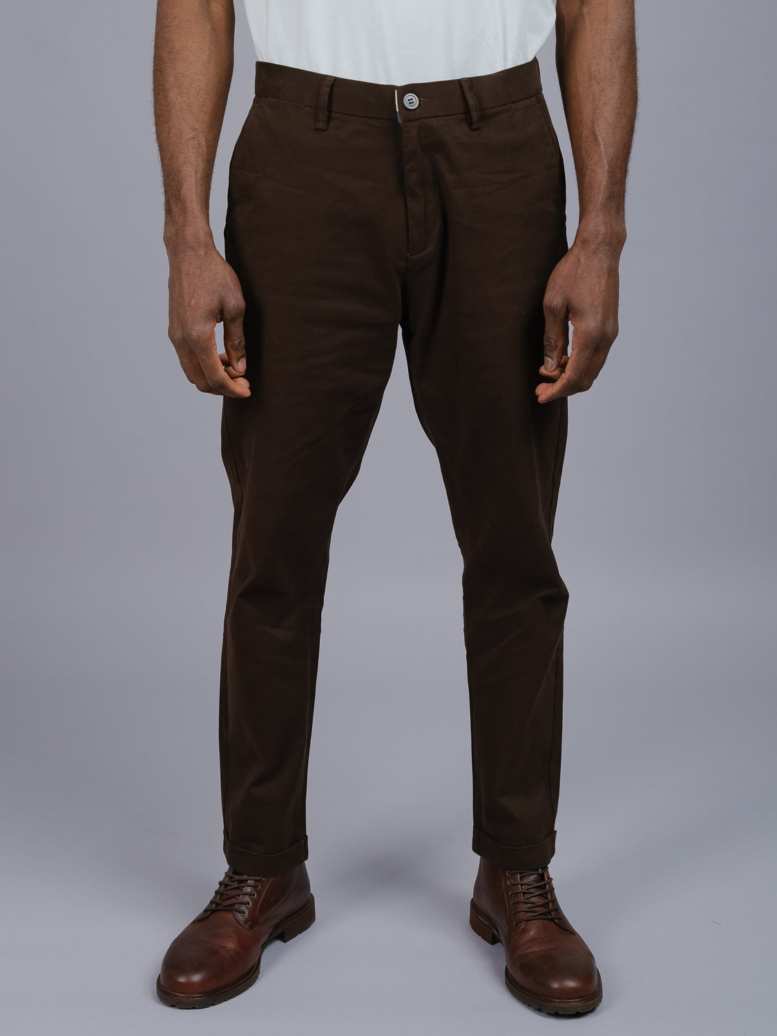 A person in a white shirt and relaxed fit ID Menswear Java Chino leather shoes stands against a plain backdrop, spotlighting the lower half with dark brown cotton twill trousers paired elegantly with the shoes.