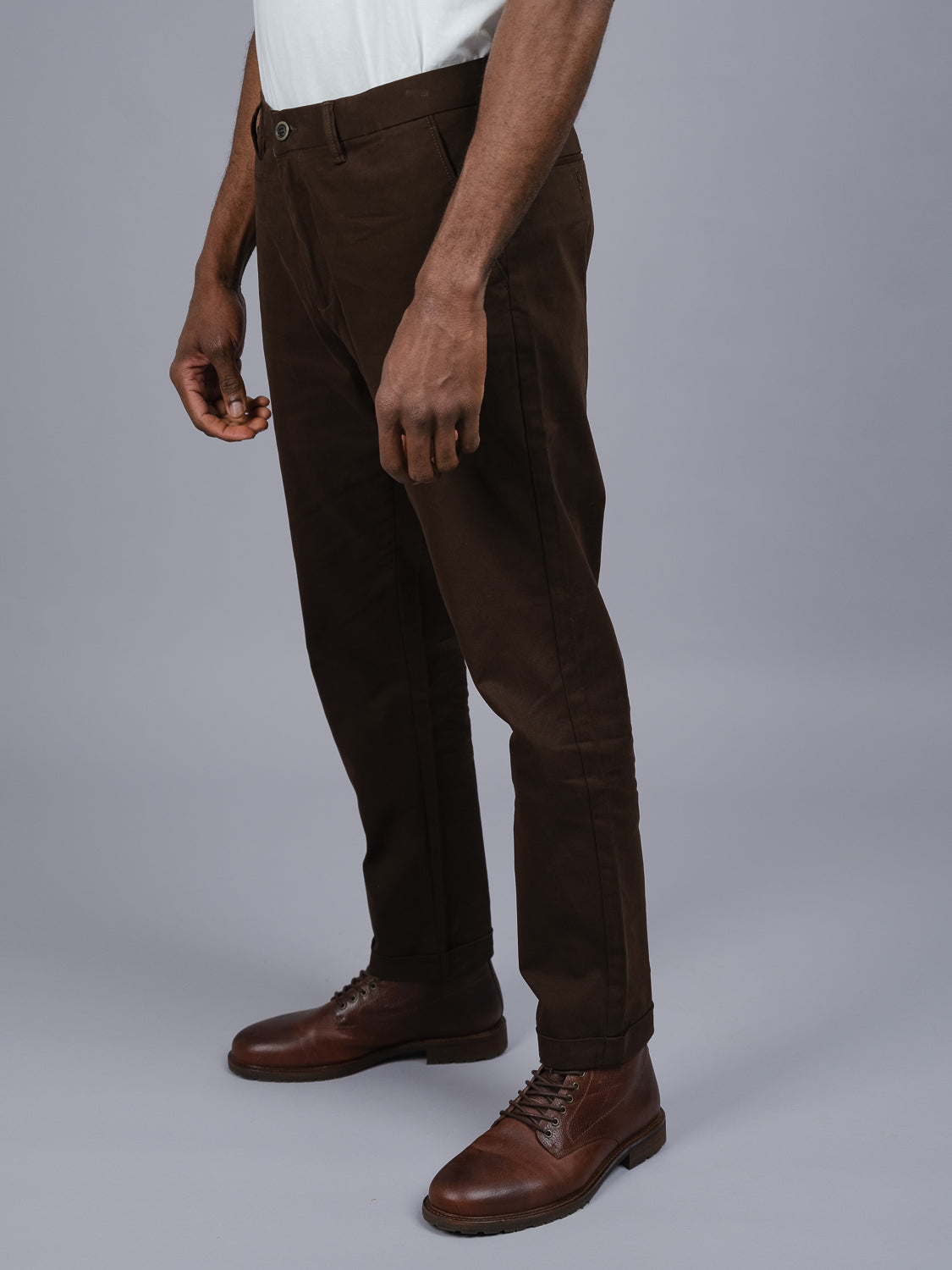 Someone wearing ID Menswears Java Chino Pant in dark brown stands against a plain gray background, paired with sleek brown leather shoes. The partially visible upper body shows arms relaxed at the sides for a casual fit.