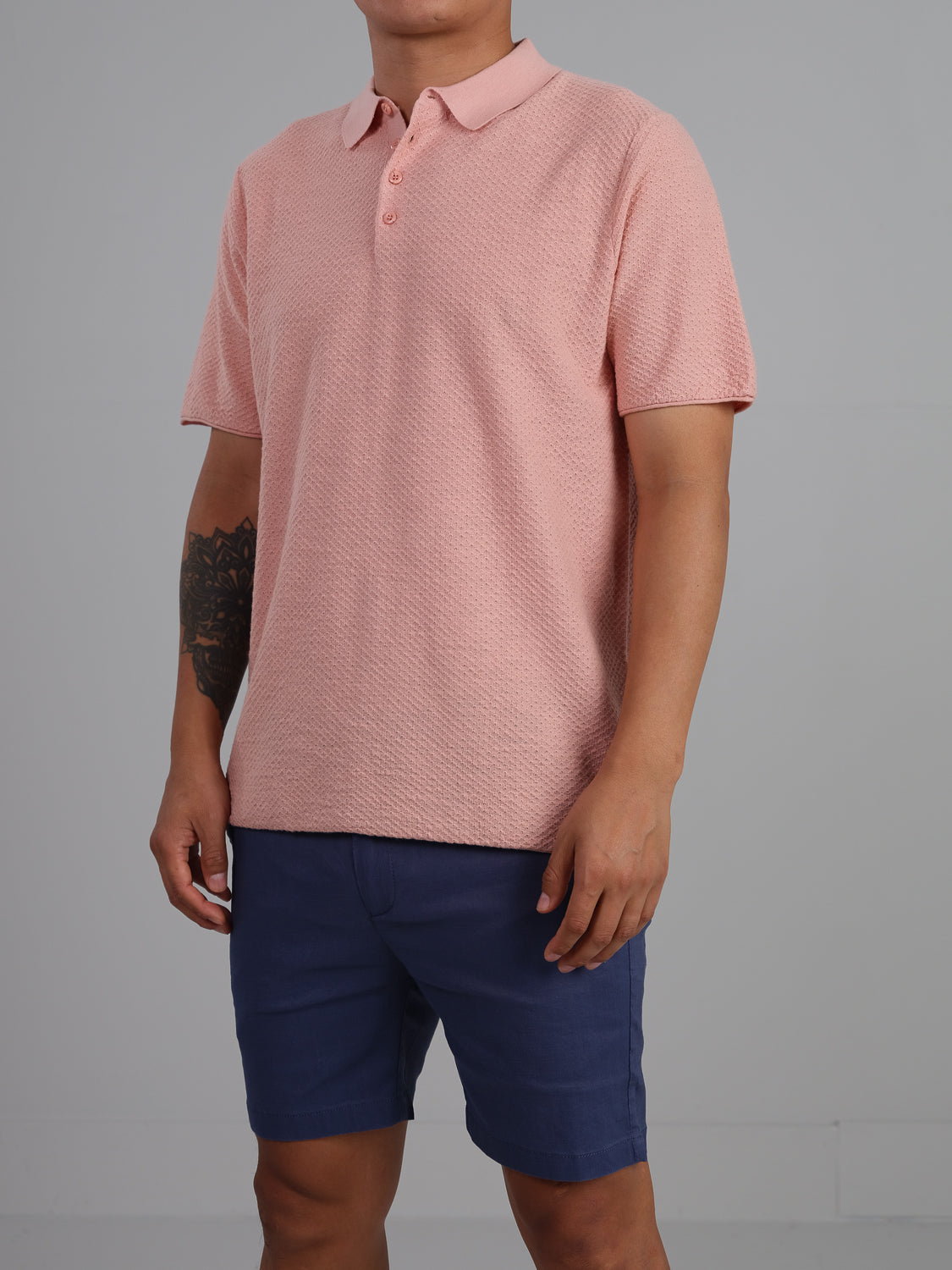 A man poses against a neutral backdrop in casual wear, featuring ID Menswears Phoenix Super Fine Cotton Knit Polo in textured pink and blue shorts, revealing a tattoo on his left forearm for a versatile stylish look.