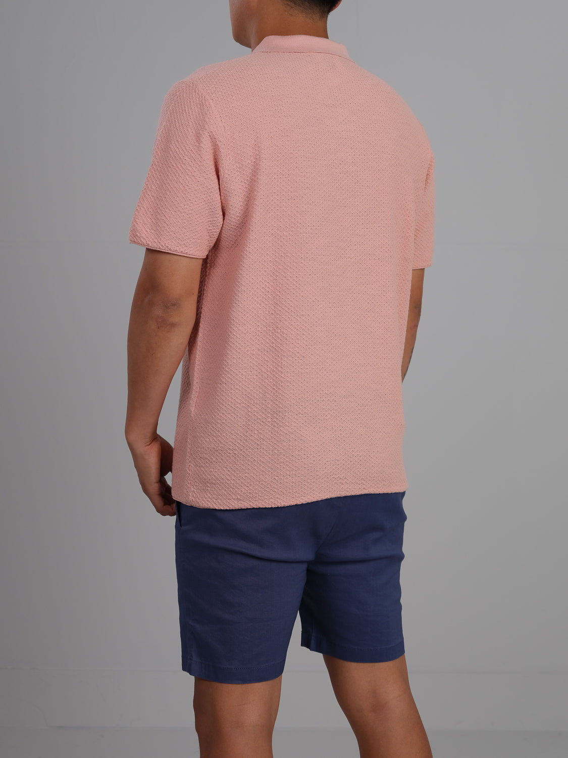 A person facing away, wearing an ID Menswear Phoenix Super Fine Cotton Knit Polo and navy blue shorts, stands against a gray background, showcasing a versatile stylish look that defines casual wear with ease.