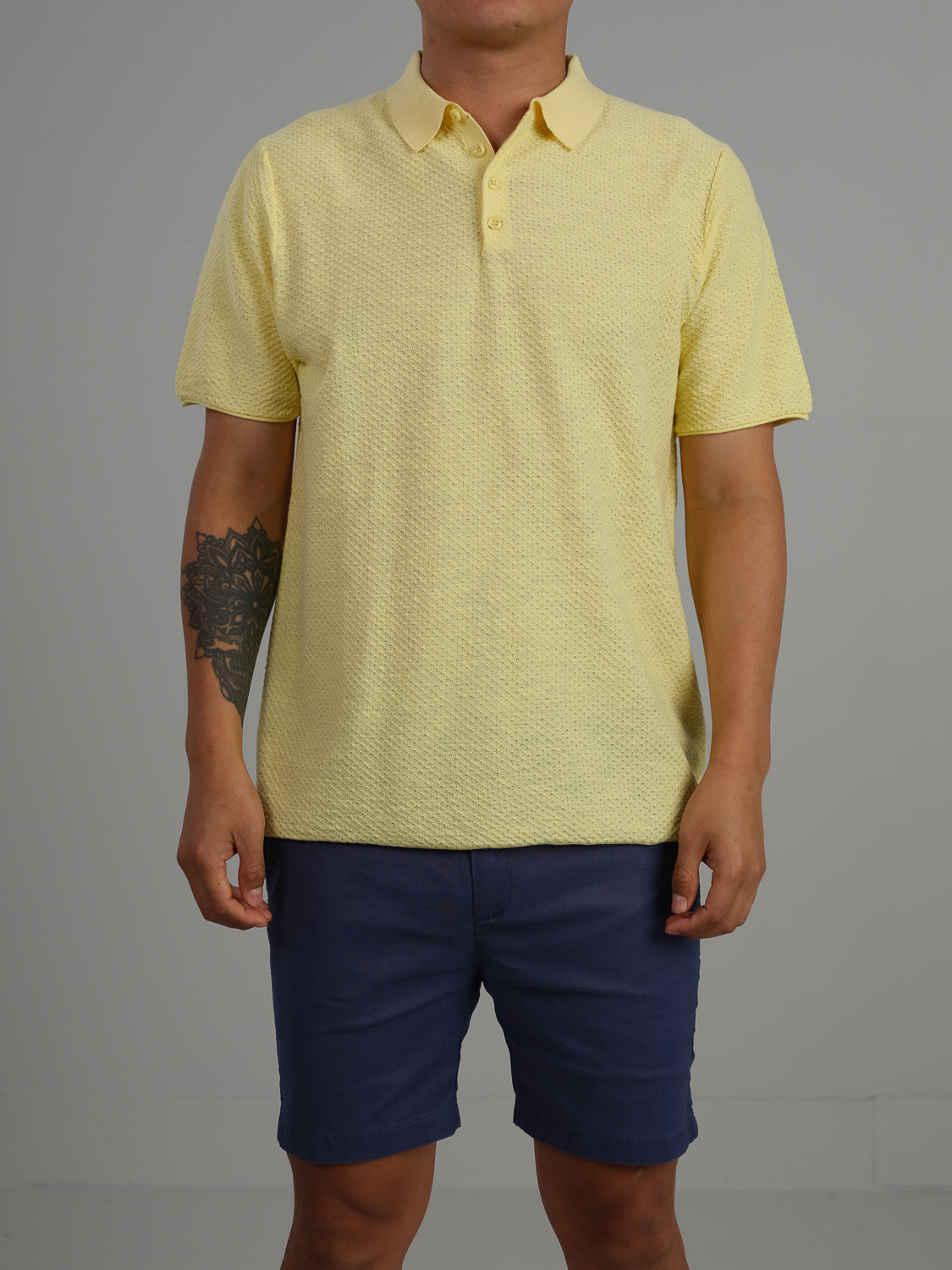 A person in a Phoenix Super Fine Cotton Knit Polo from ID Menswear, in light yellow with a subtle geometric pattern, paired with dark blue shorts, stands against a plain gray background. The polo complements the tattoo on their left forearm, creating a stylish look ideal for casual wear.