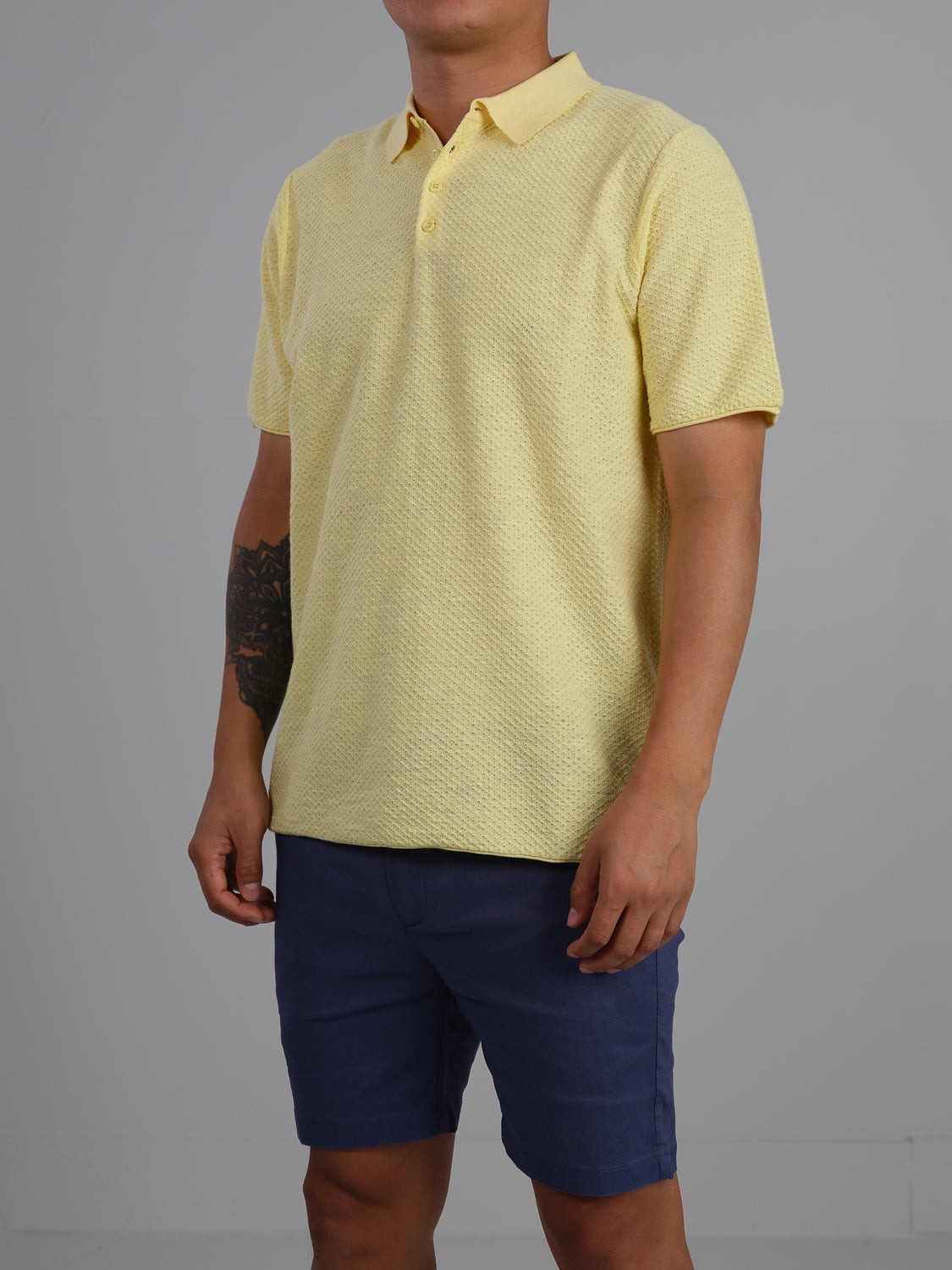 A person sports a chic and versatile look with the textured Phoenix Super Fine Cotton Knit Polo from ID Menswear in yellow, paired with dark blue shorts, against a plain gray background, displaying a tattoo on their left forearm.