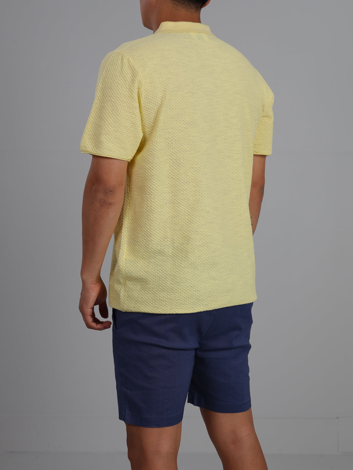 Wearing an ID Menswear Phoenix Super Fine Cotton Knit Polo in yellow and blue shorts, a person projects a versatile, stylish look as they stand with their back to the camera against a plain gray background.