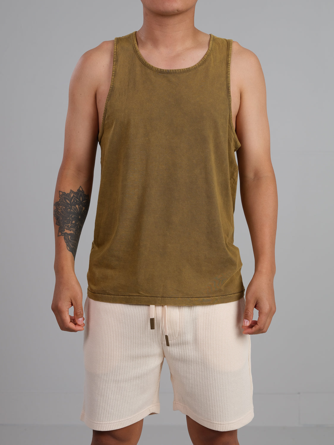 Wearing an ID Menswear Settler Snow Vintage Wash Tank Top and light shorts, the tattooed person poses against a plain gray background, with their head cropped out of the image.